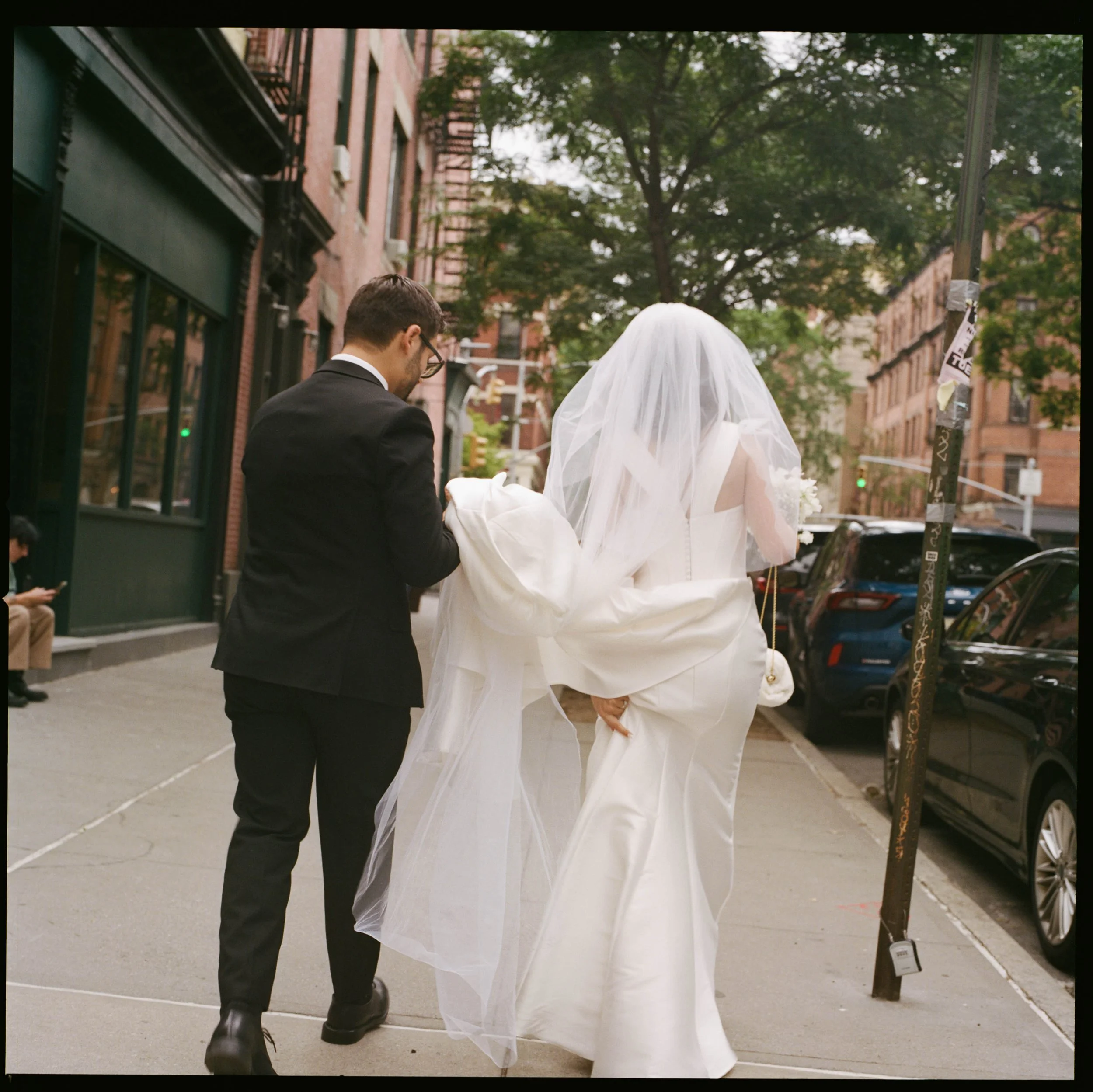 A man in a black suit helping a woman in a white wedding dress and veil walk on a city sidewalk.