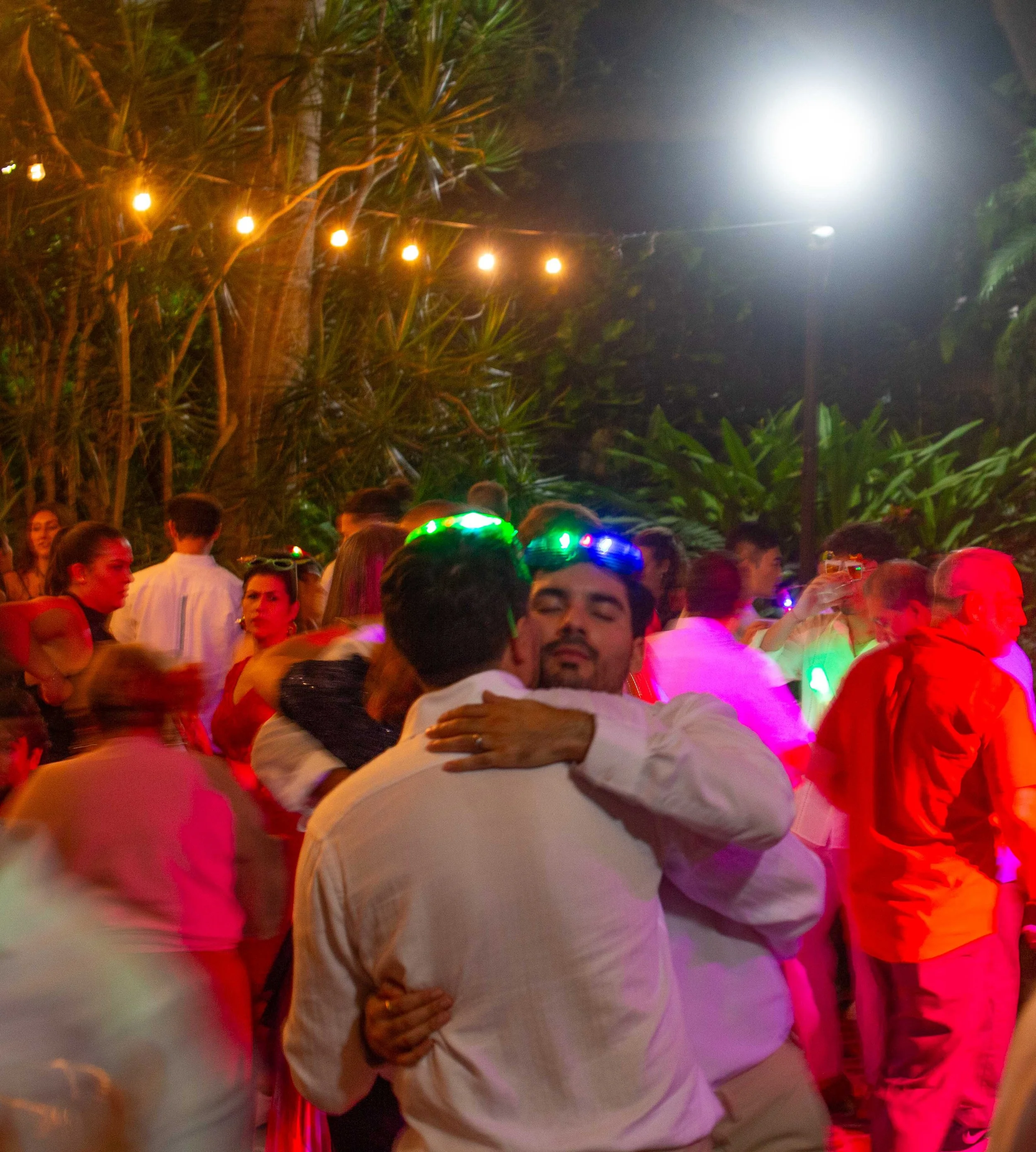 People dancing and hugging at an outdoor party at night, decorated with string lights and lush greenery.