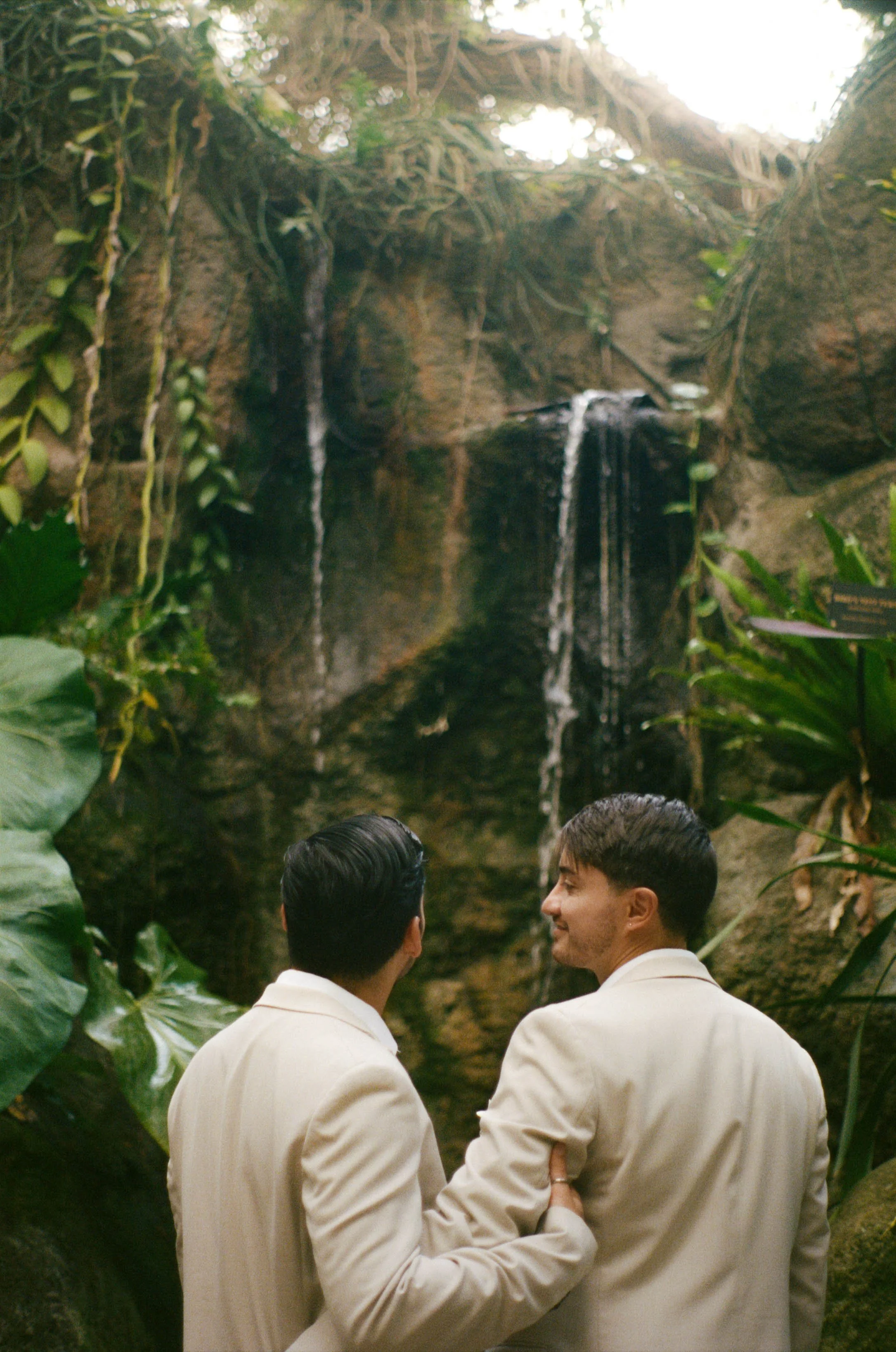 Two men in beige suits standing close together, smiling, with a waterfall and lush greenery in the background.