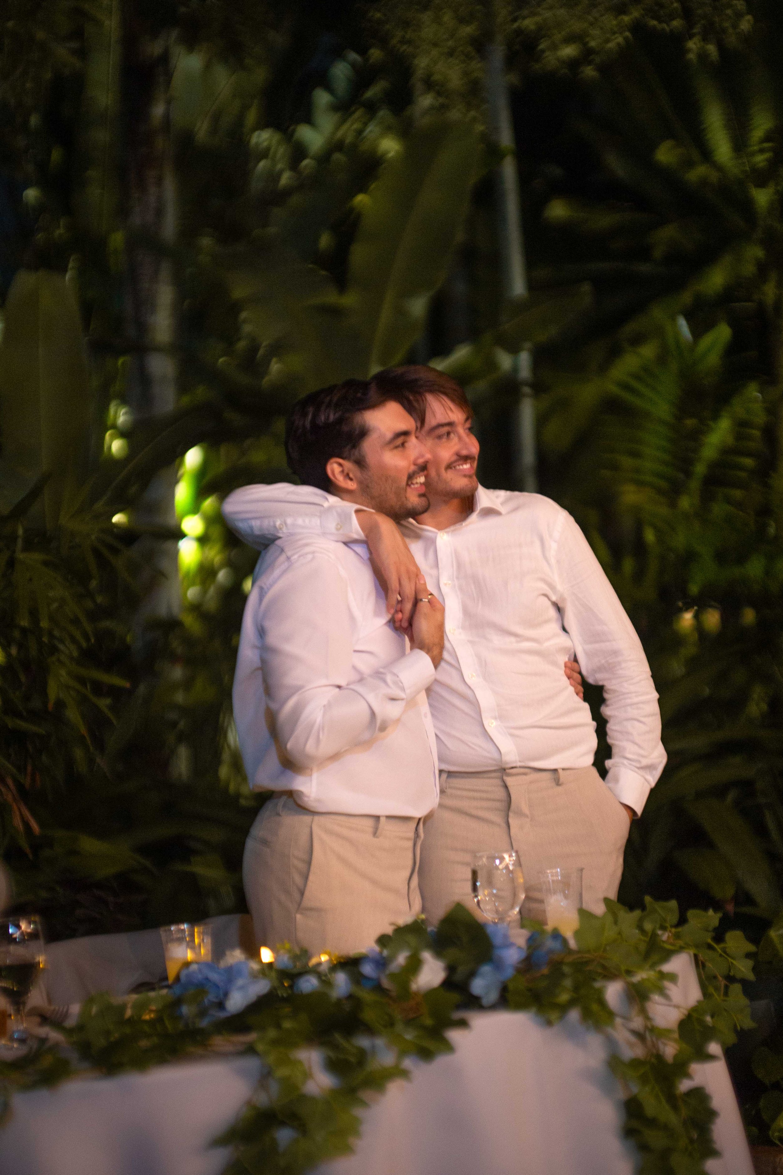 Two men dressed in white shirts and beige pants are smiling and standing close together at a decorated table, with one man’s arm around the other's shoulders. The background is filled with large green plants, suggesting an outdoor or tropical setting