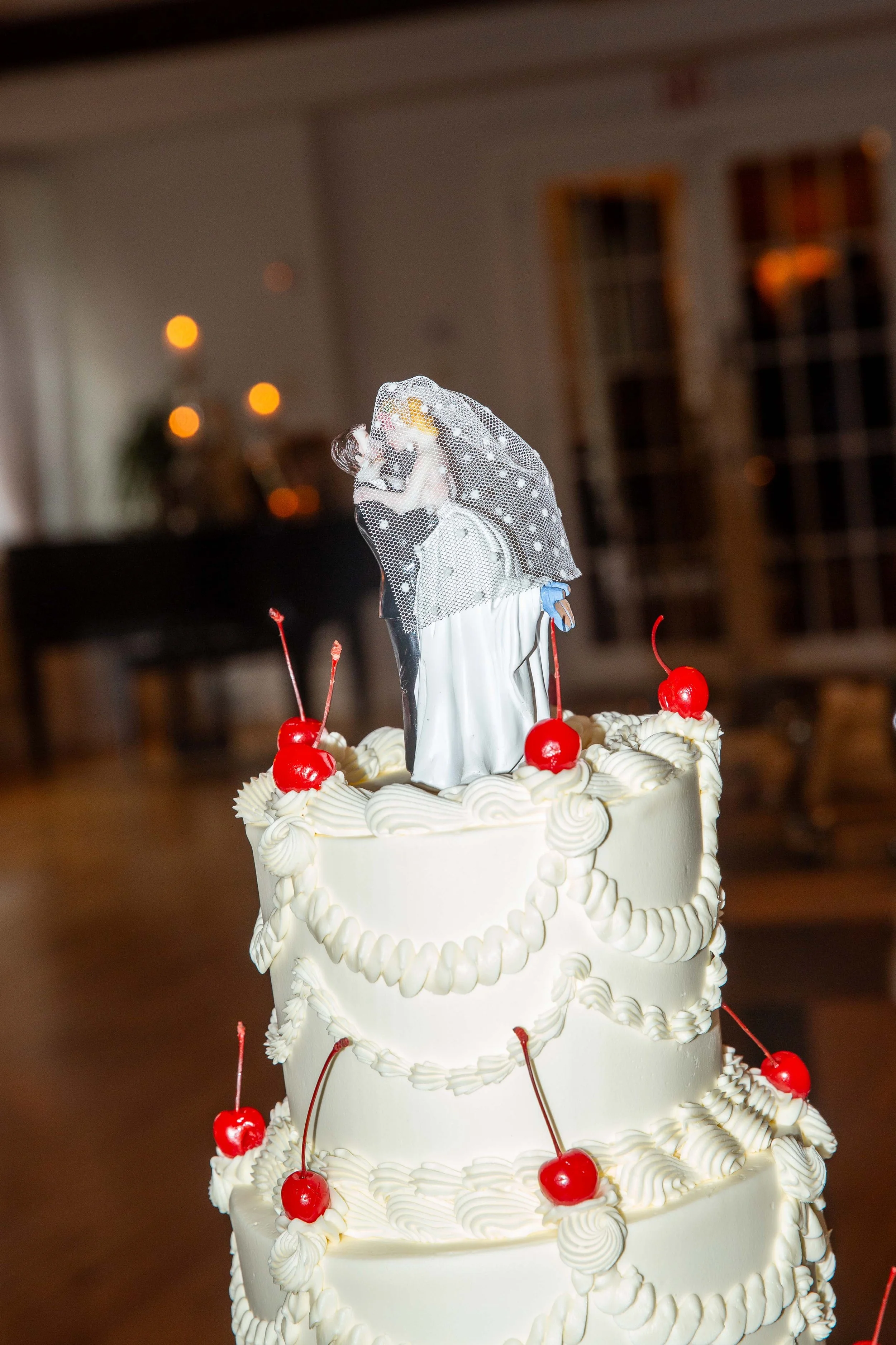 Three-tier wedding cake with red cherries on top of each layer, white icing with piped rosettes and garlands, topped with a figurine of a bride and groom in wedding attire.