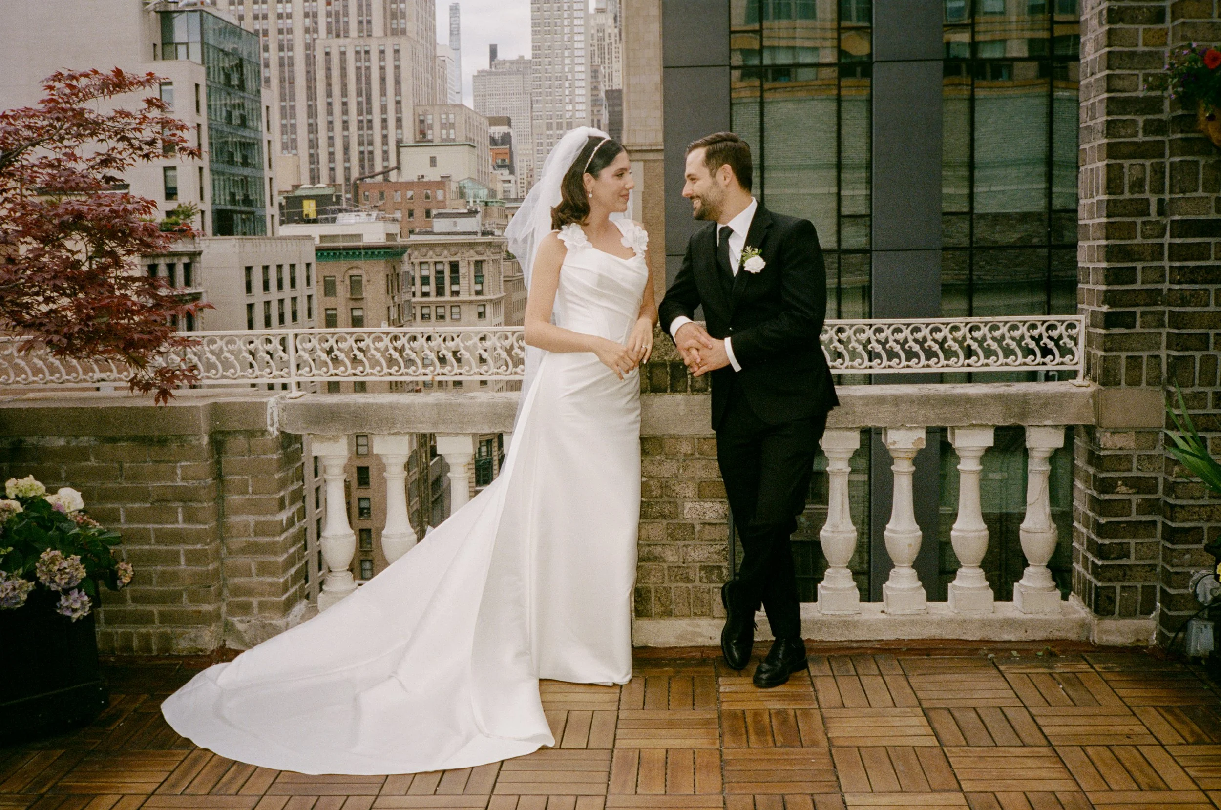 A bride in a white wedding gown and veil and a groom in a black suit and tie standing on a balcony with a cityscape background, smiling at each other.