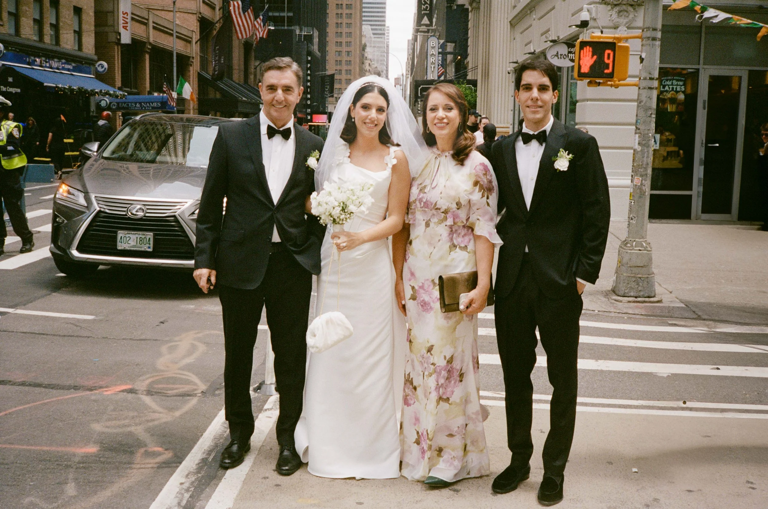 A wedding celebration in an urban setting, featuring a bride in a white dress holding a bouquet, standing next to three people dressed in formal attire, with a city street, cars, and buildings in the background.
