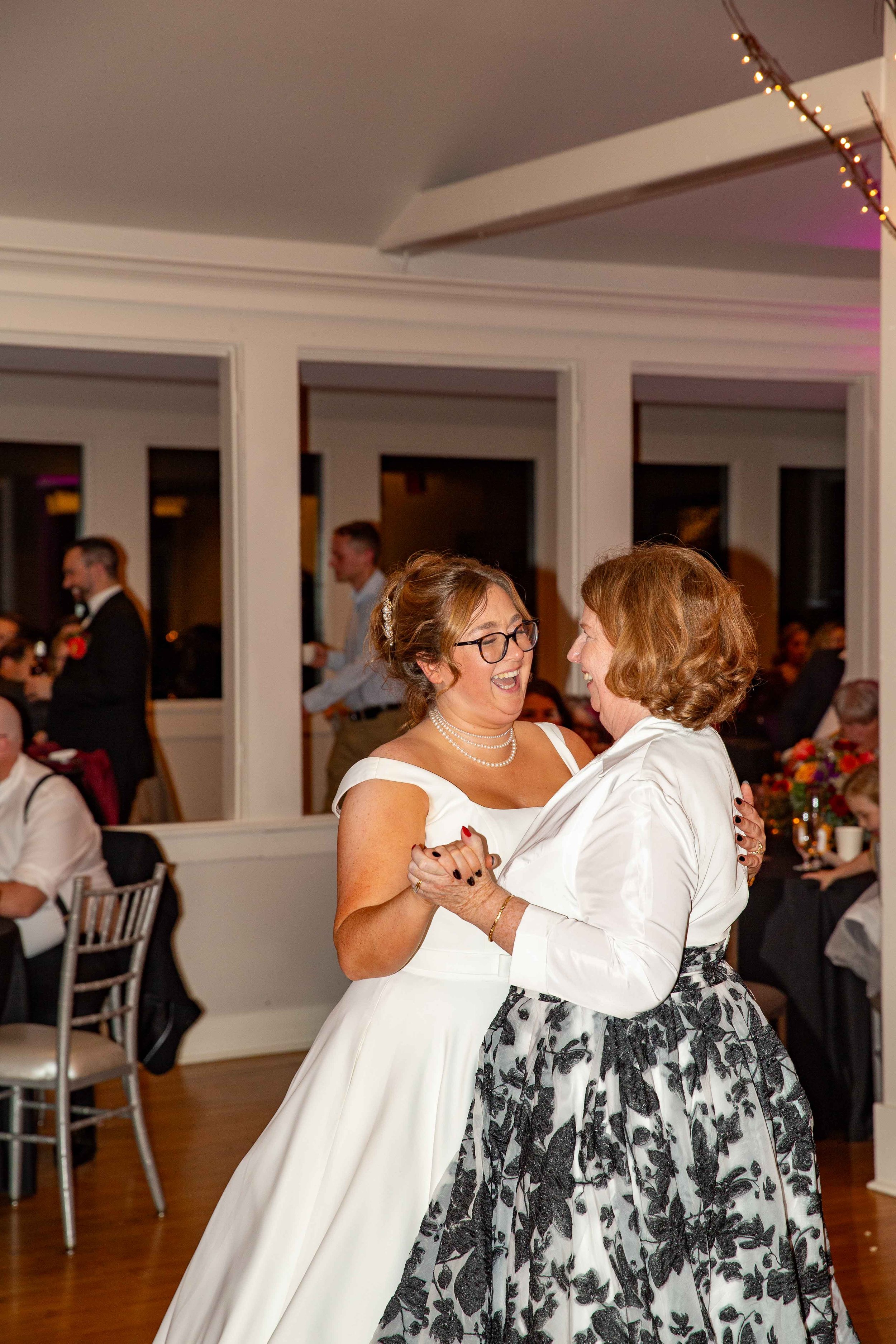 Two women dancing and smiling at a wedding reception with other guests in the background.