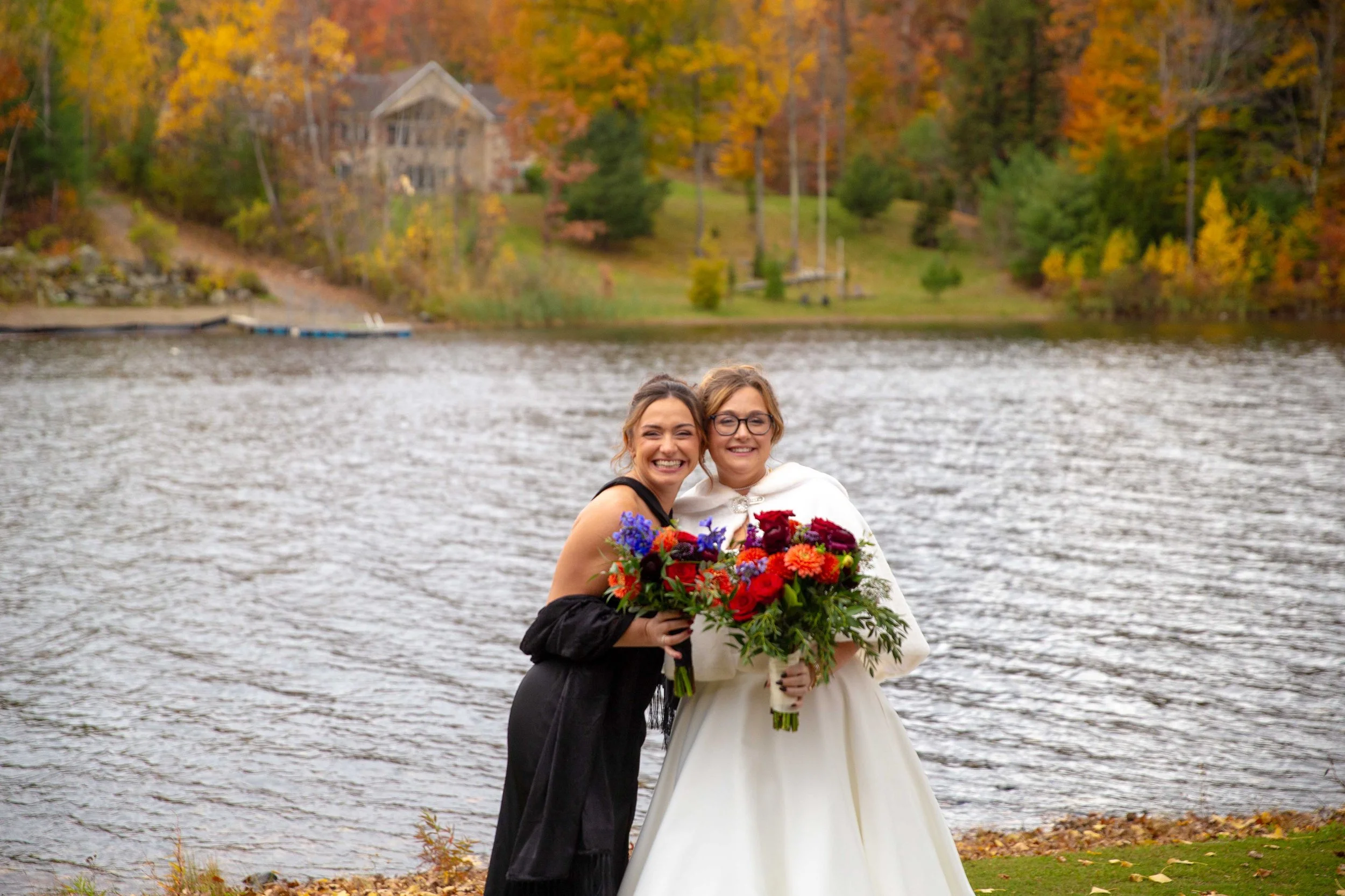 Two women standing by a lake, smiling and holding bouquets of colorful flowers, with fall foliage and a house in the background.