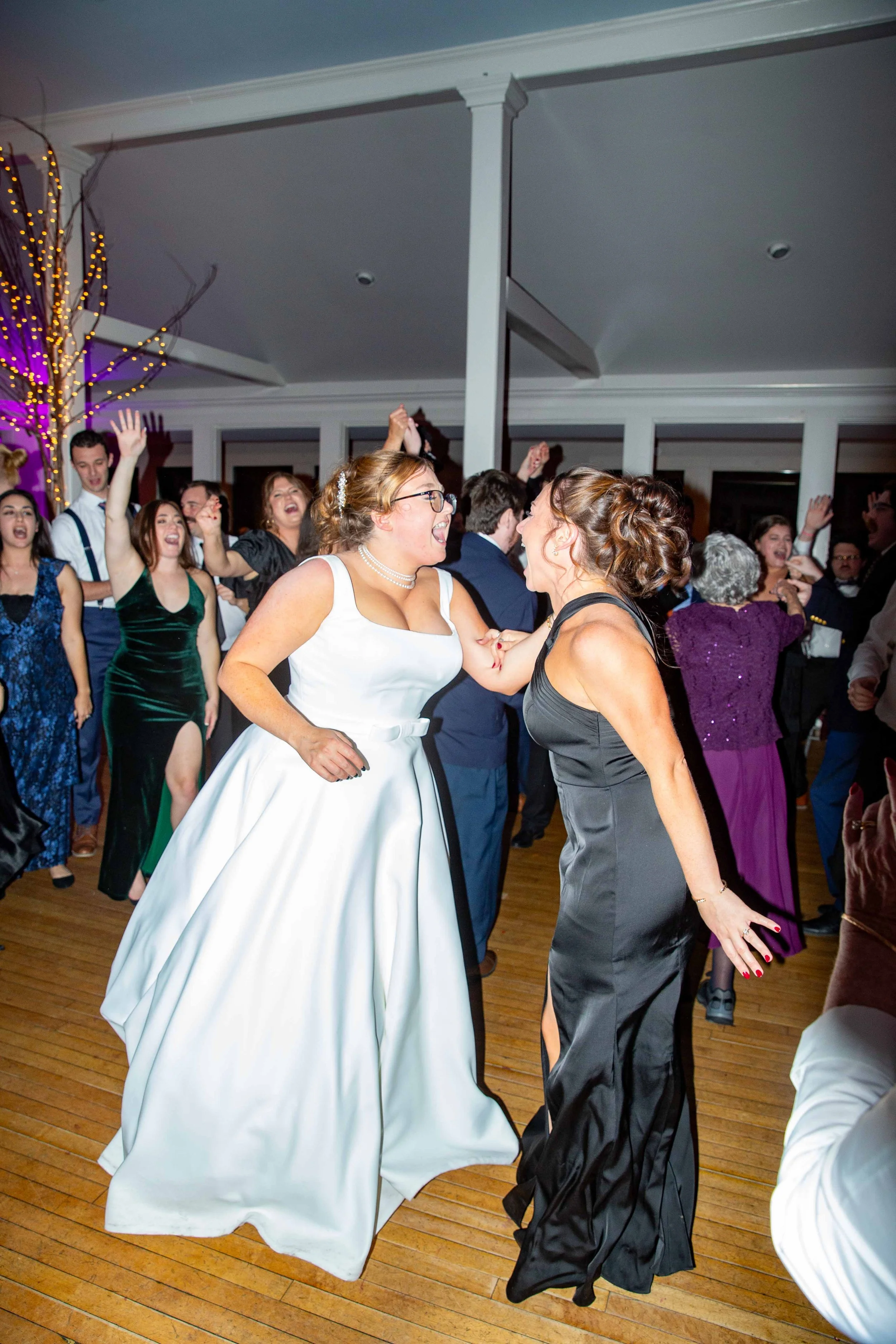 Two women, one in a white wedding dress and the other in a black dress, are happily dancing and laughing together at a wedding reception with other guests dancing in the background.