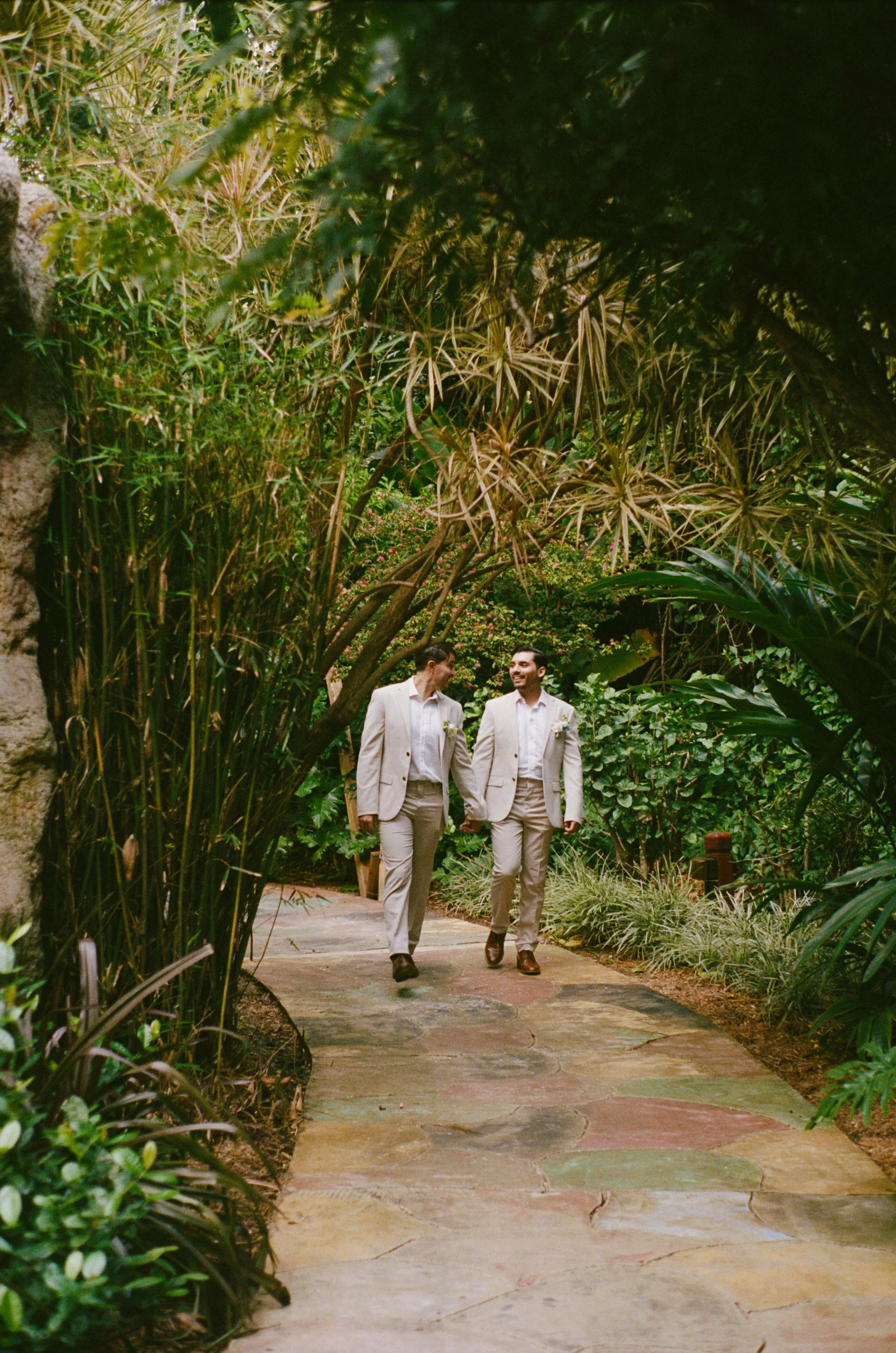 Two men in beige suits walking hand in hand on a garden path, surrounded by lush green plants and trees.