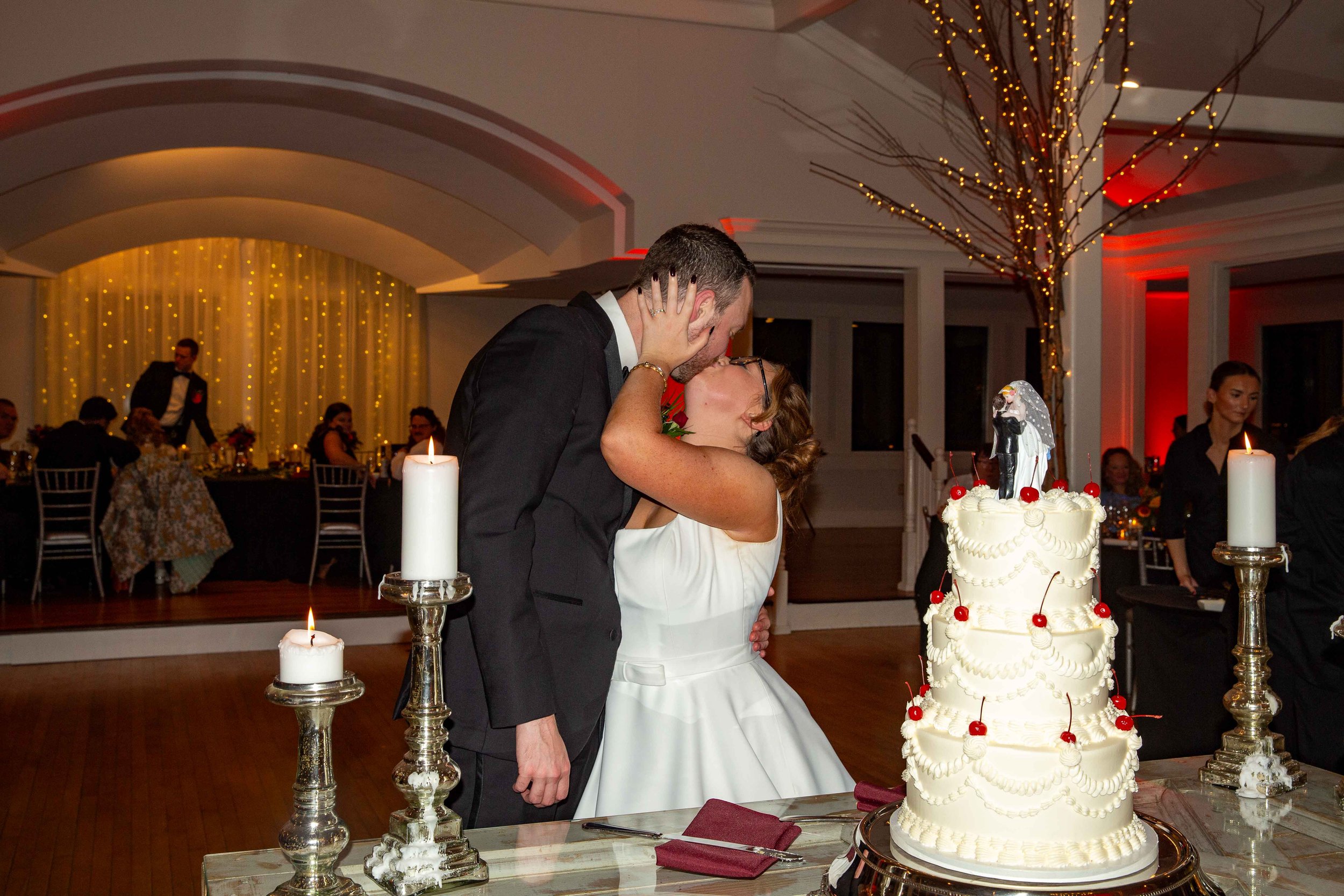 A bride and groom kiss at their wedding reception, with a wedding cake and candles on the table in front of them, and guests seated in the background.