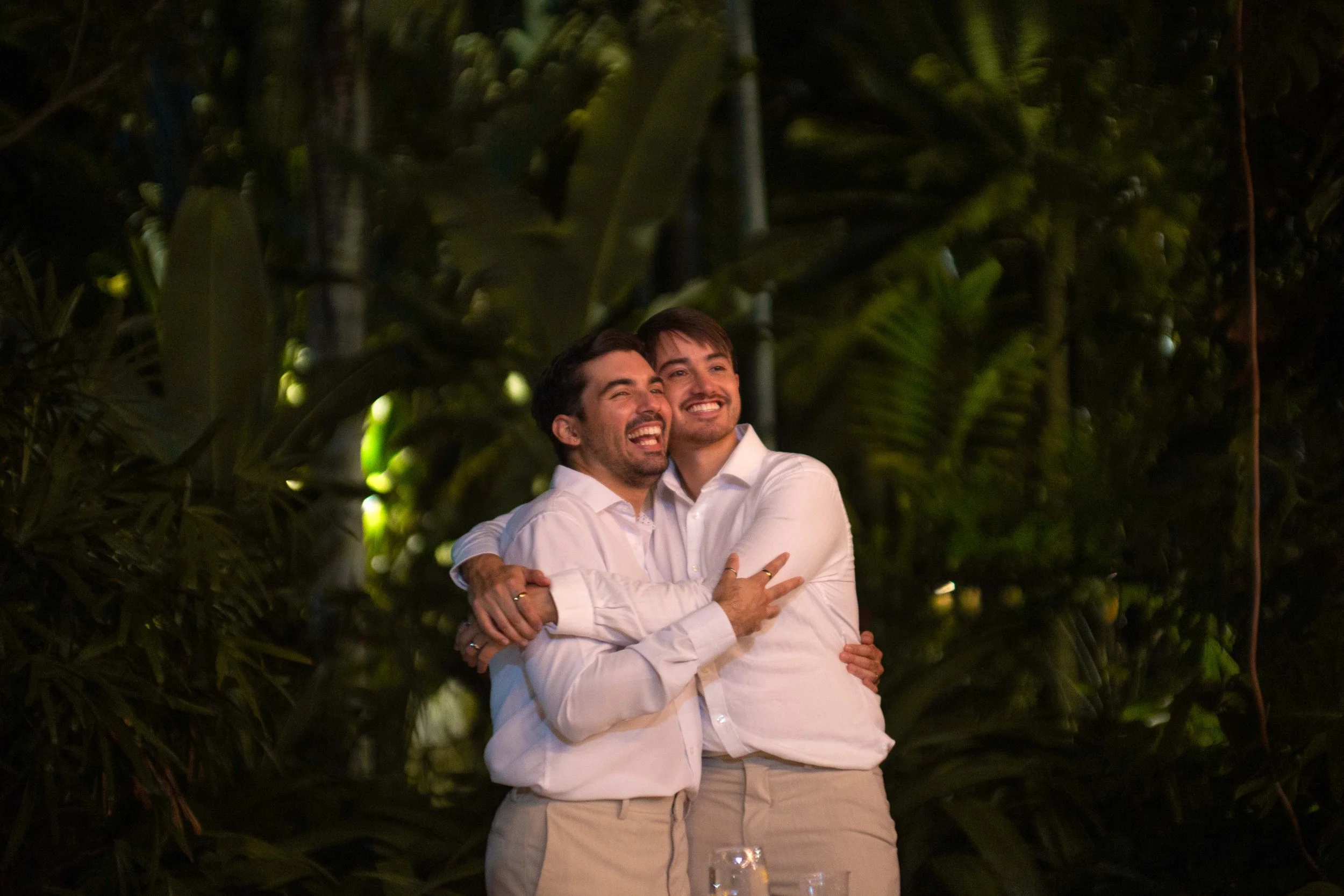 Two men in white shirts hugging and smiling in an outdoor setting at night, surrounded by green foliage.