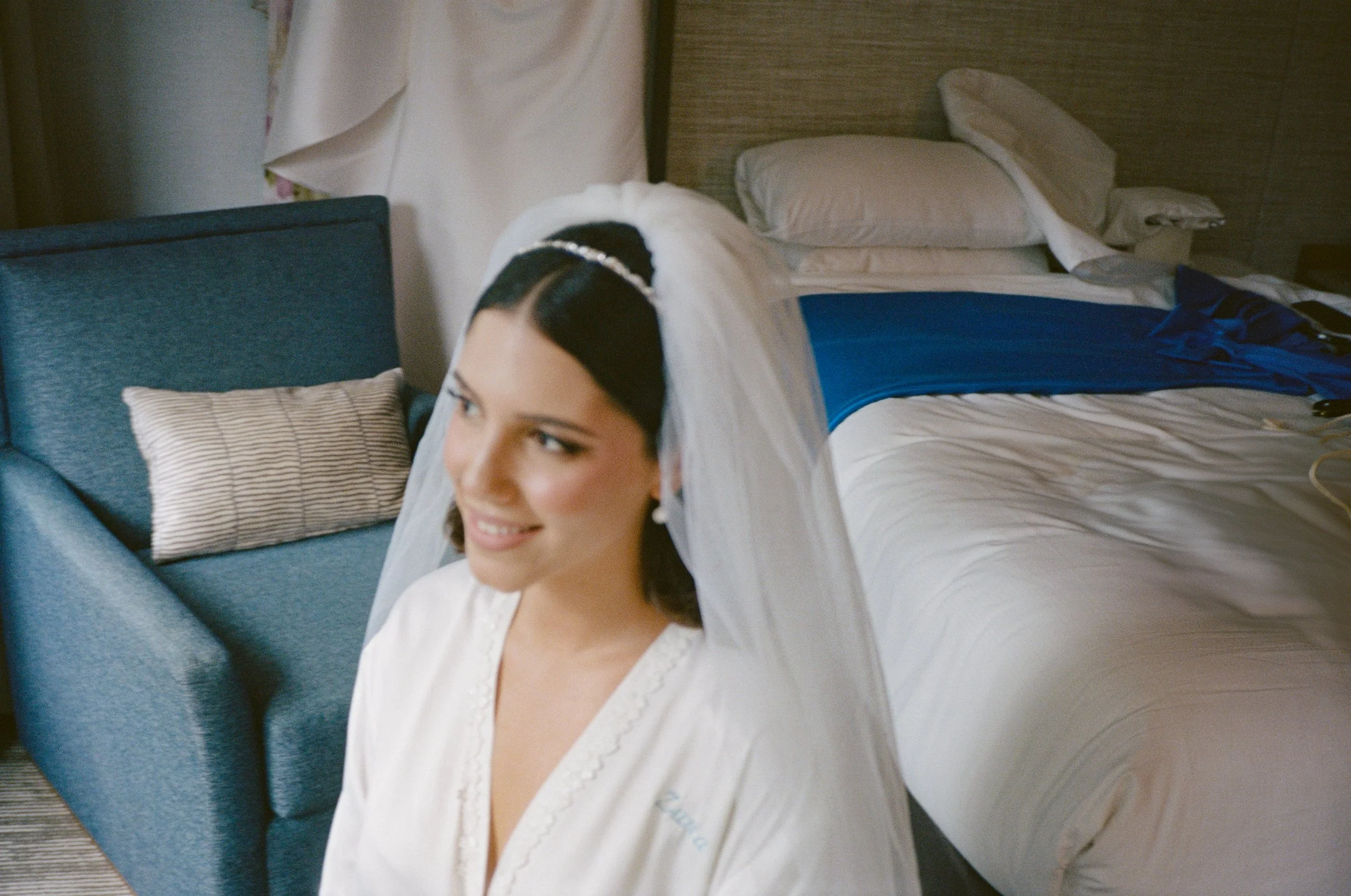 A smiling woman dressed in a wedding gown, sitting in a hotel room near a bed with rumpled linens and pillows.