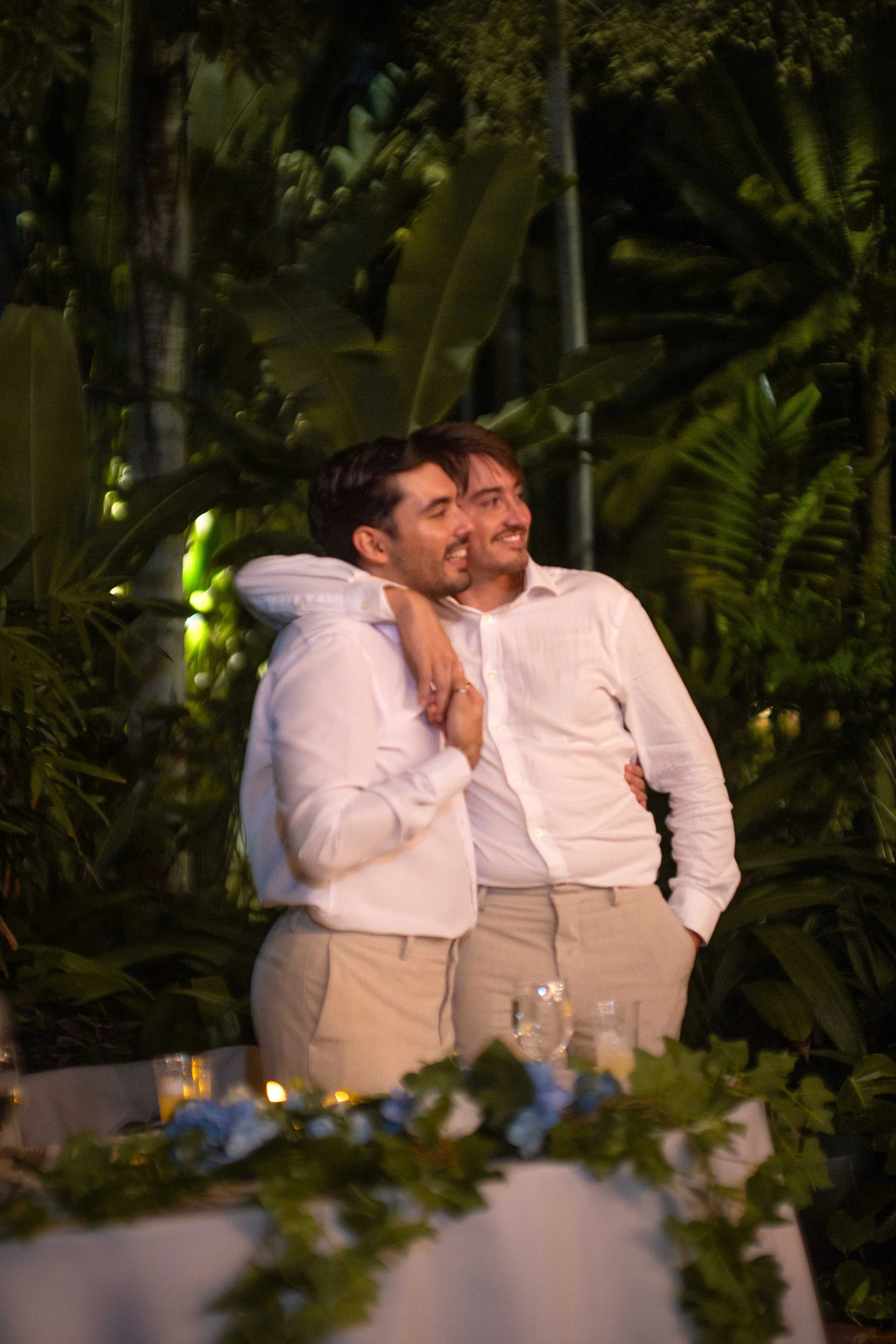 Two men in white shirts and beige pants standing close together with one man's arm around the other's shoulders, smiling, in an outdoor setting with large green tropical plants in the background.