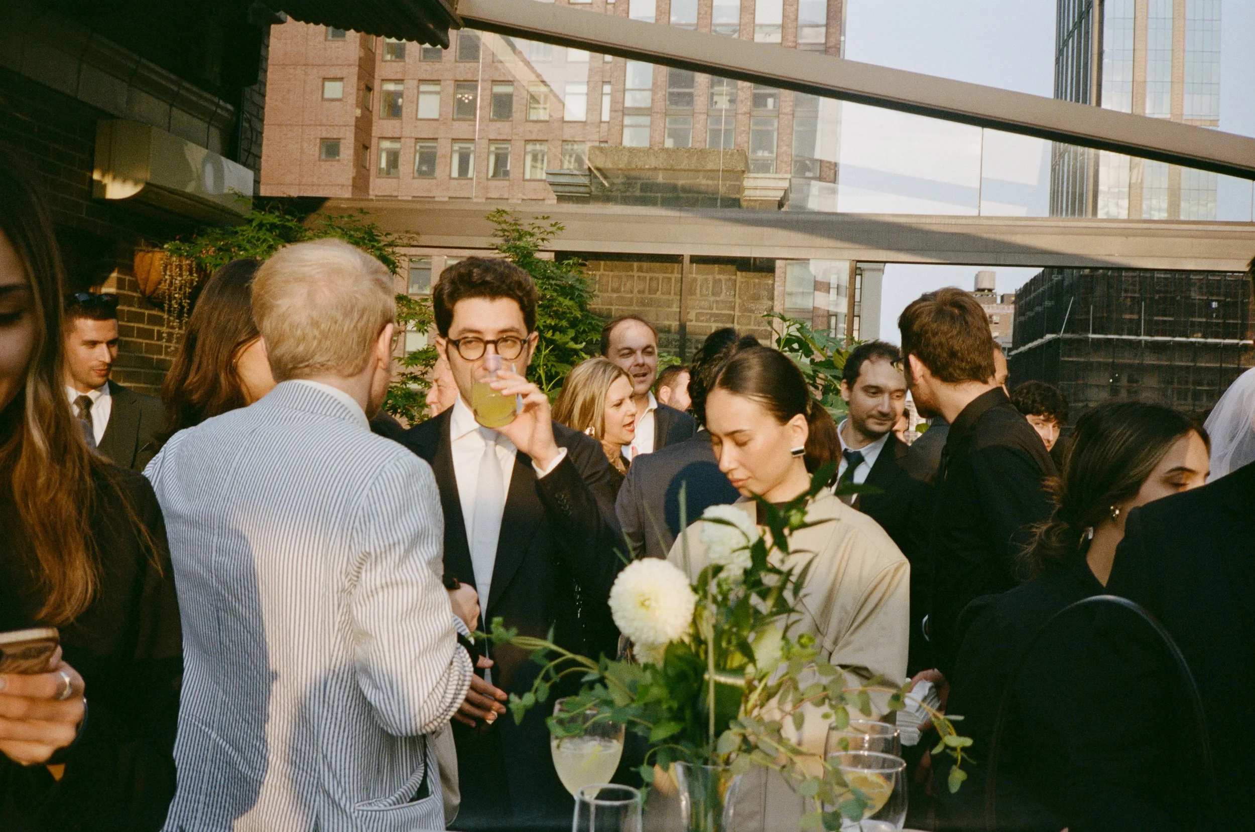 A group of people at a rooftop social gathering, some talking, one person drinking a beverage, with city buildings in the background and floral centerpieces on tables.