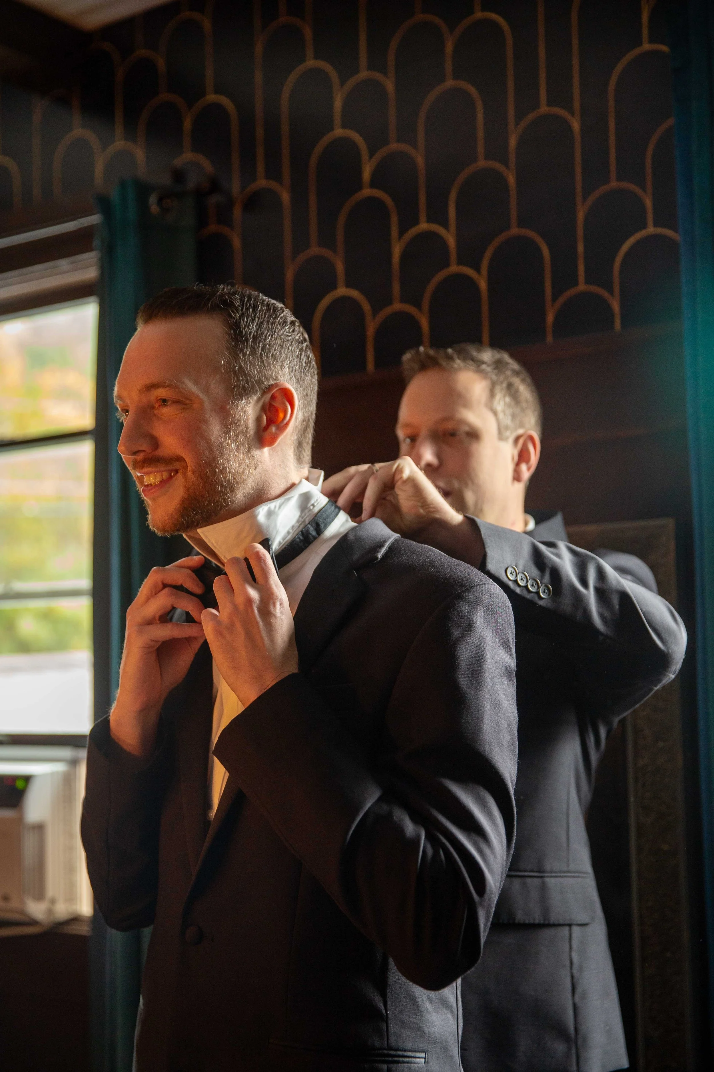 Two men in suits helping each other get ready, one adjusting the collar of the other's shirt.
