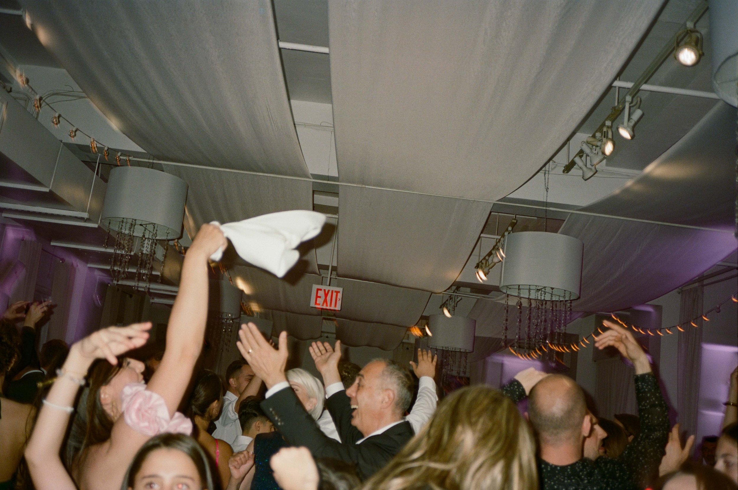 People dancing and celebrating at a party with their hands up, some clapping, in a decorated indoor venue with hanging lights and ceiling panels.