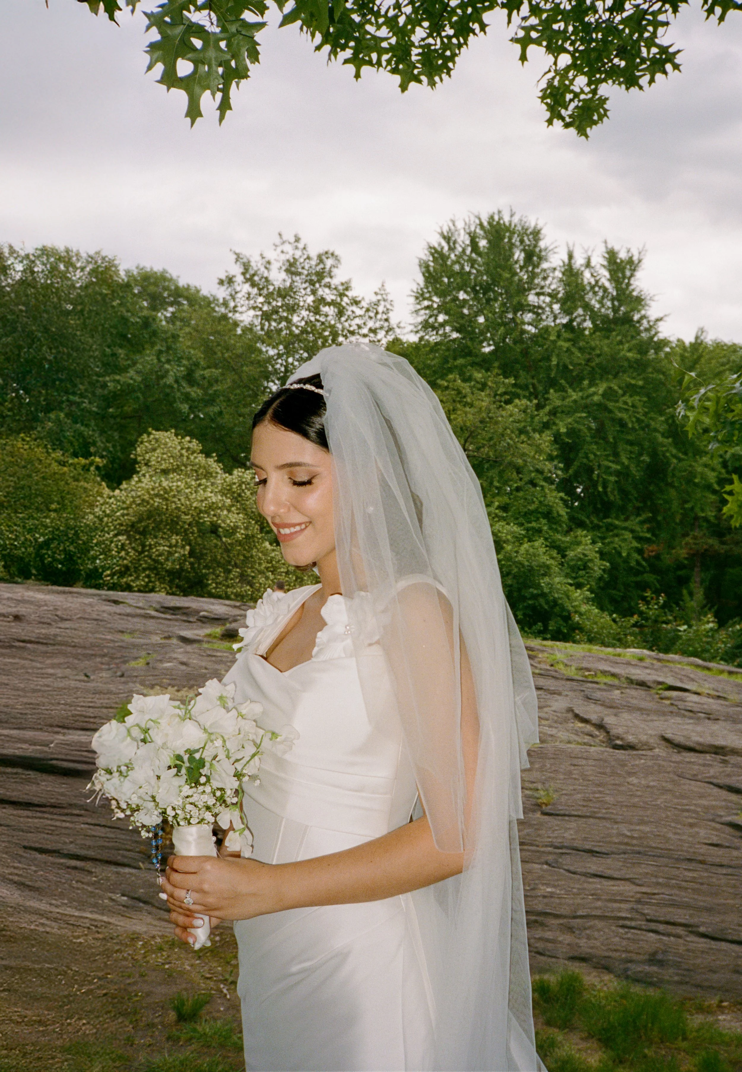 A bride in a white wedding dress holding a bouquet of white flowers, standing outdoors with trees and a cloudy sky in the background.