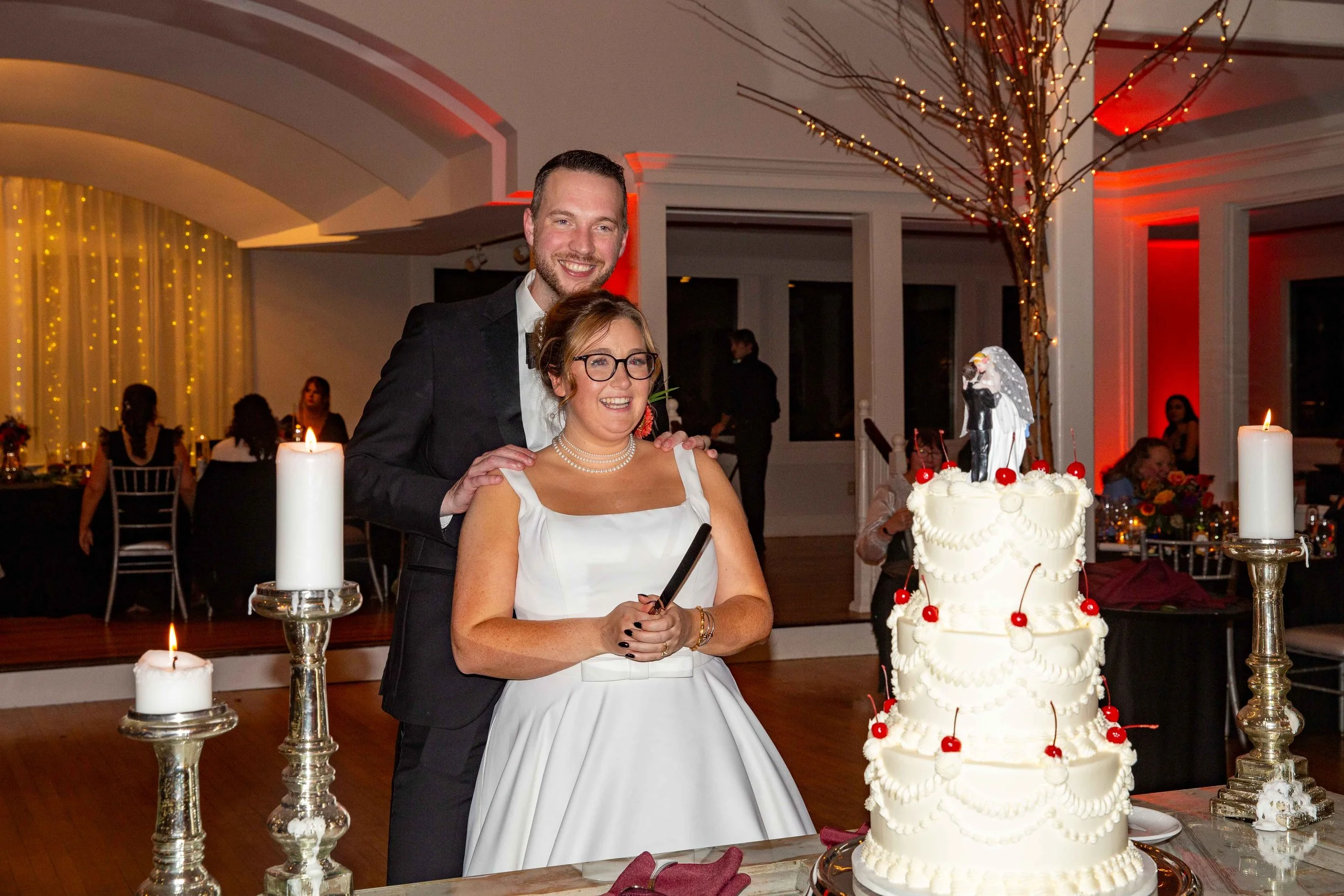 A bride and groom at their wedding celebration, standing behind a tiered wedding cake decorated with cherries and a bride and groom cake topper, with candles on tall silver holders and guests seated in the background.