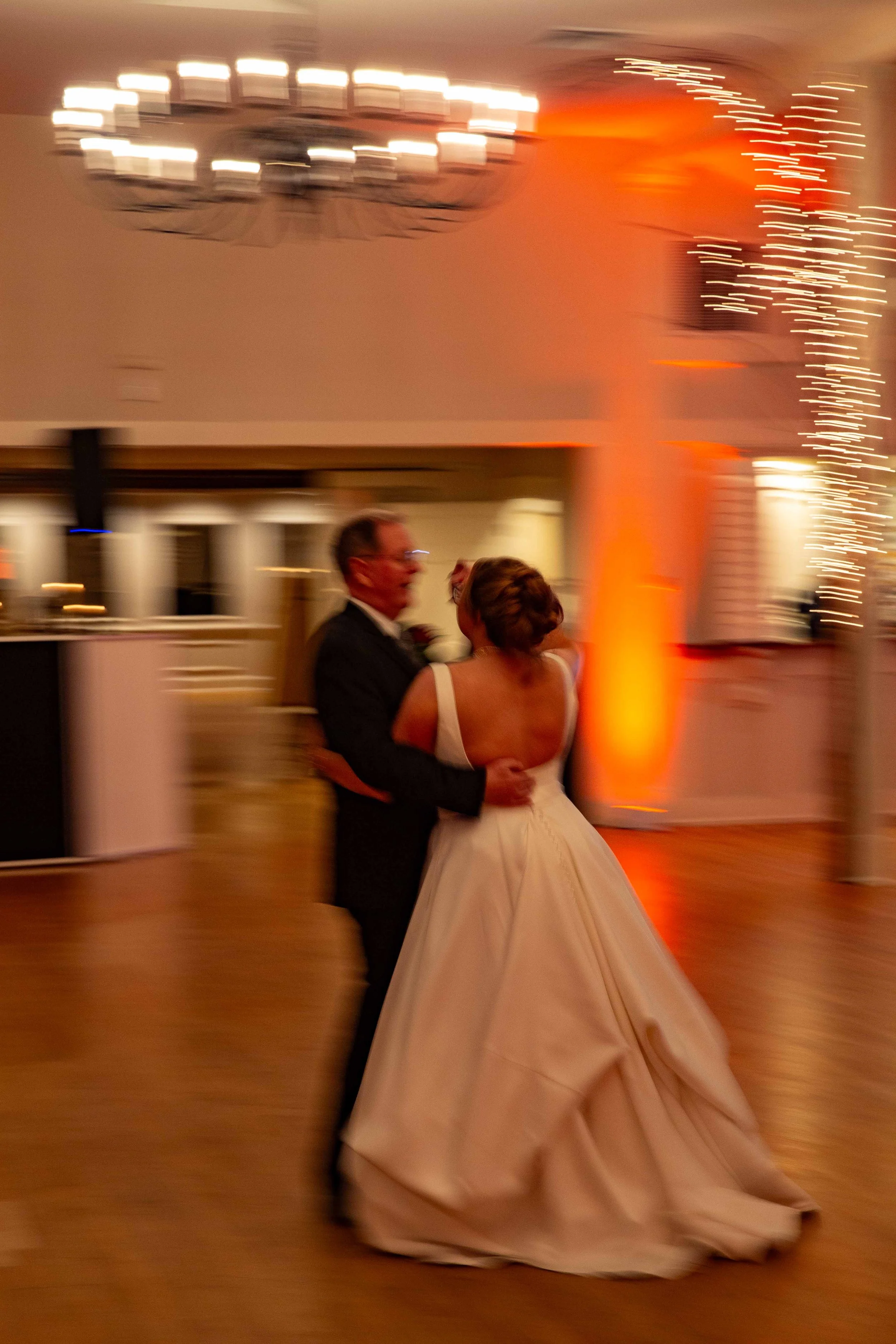 A couple dancing at a wedding reception, with the bride in a white gown and the groom in a black tuxedo, in a warmly lit room with festive lighting and a chandelier.