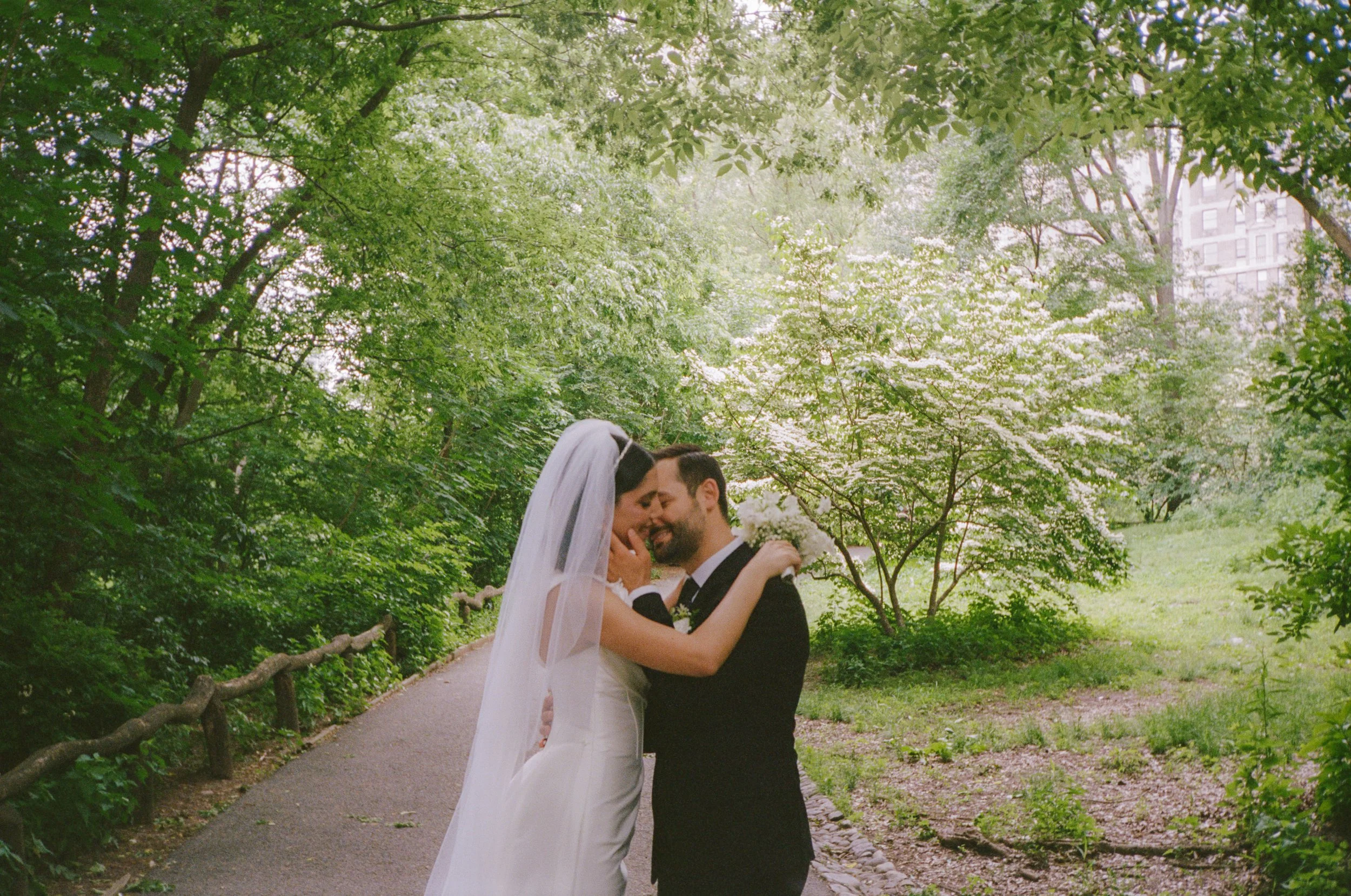 A bride and groom embrace in a lush, green park with trees and blooming flowers, smiling and touching foreheads.