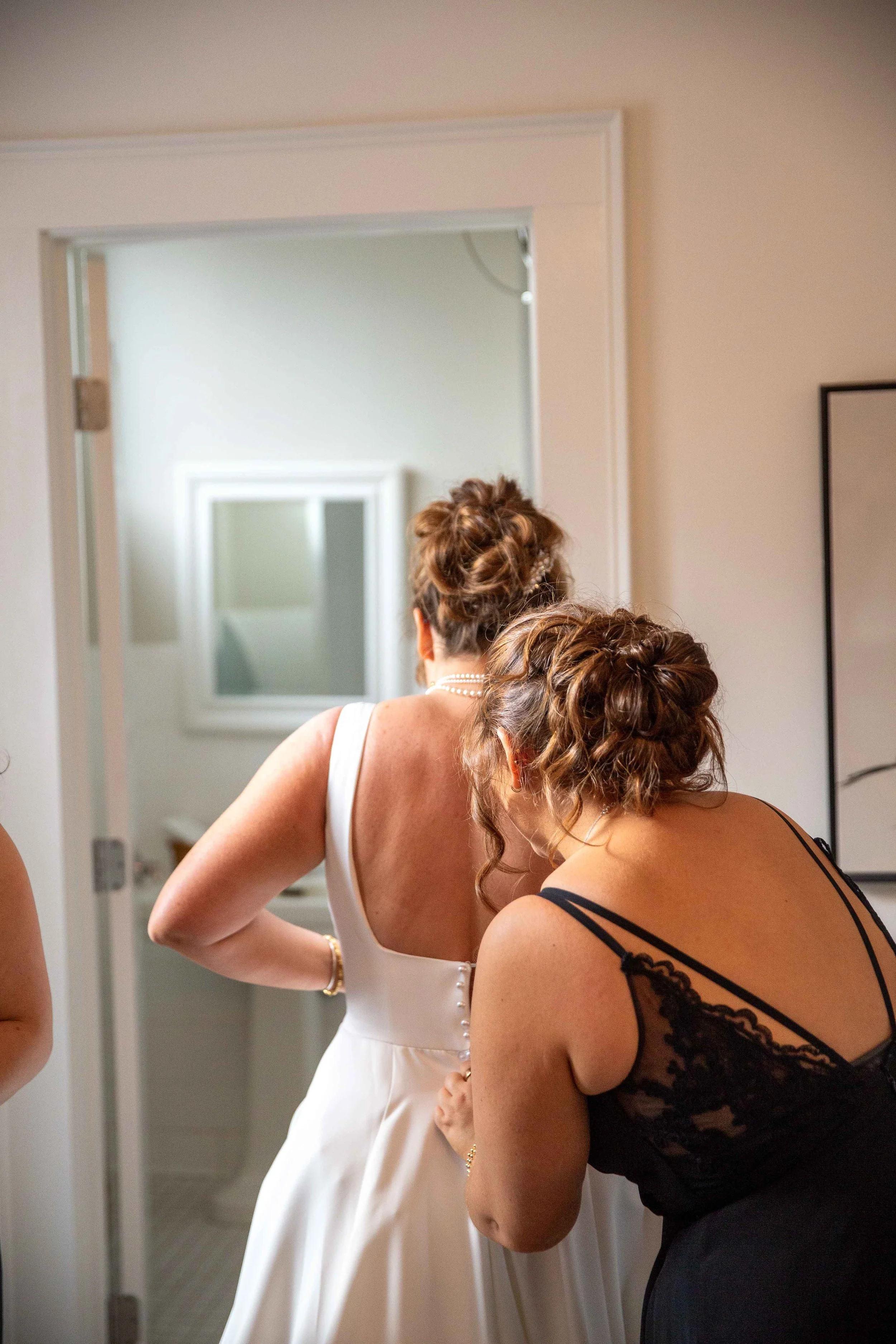Two women preparing for a wedding, one in a white wedding dress and the other in a black lace dress, adjusting the bride's gown.