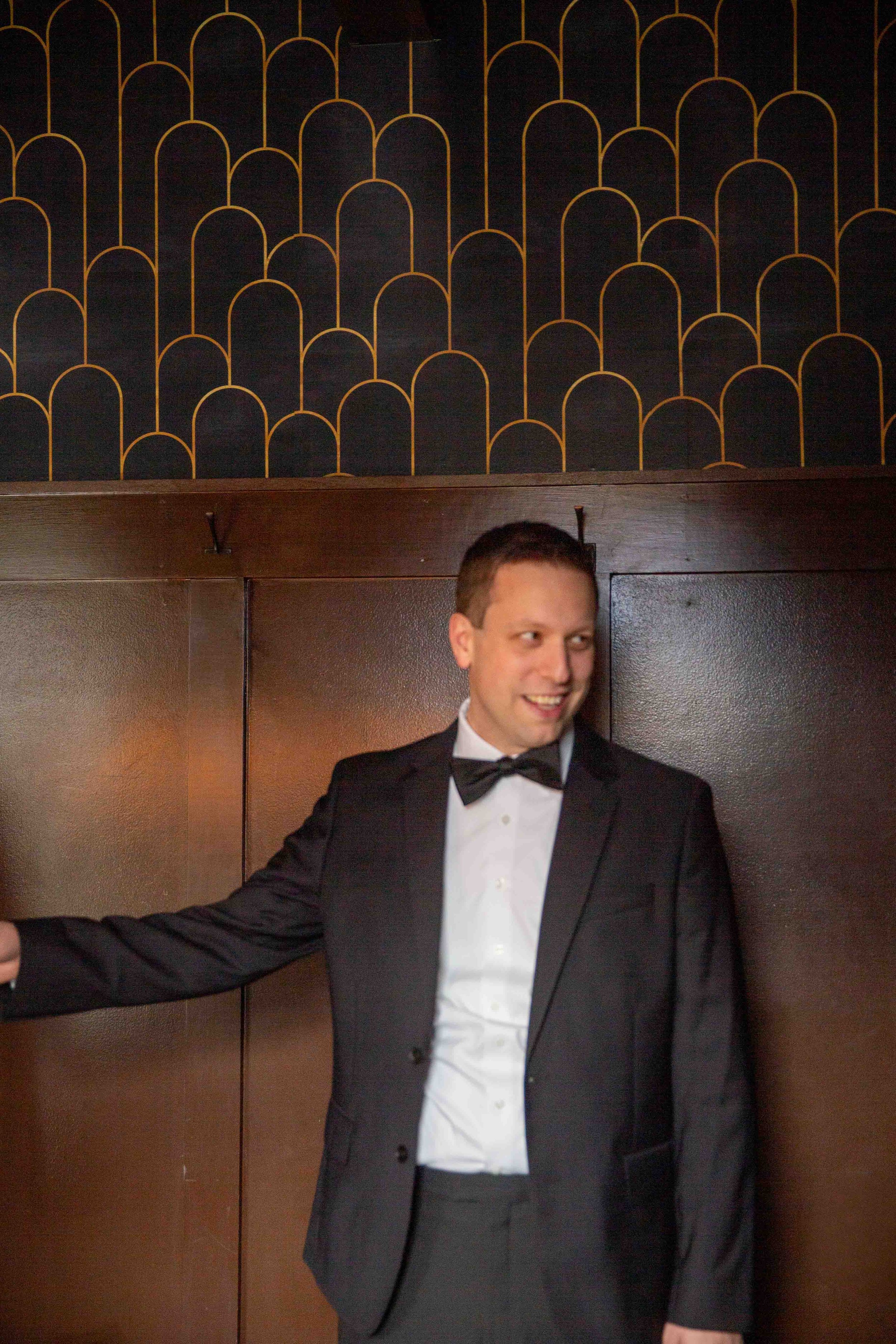 A man dressed in a tuxedo with a bow tie standing against a dark wooden wall with art deco patterned wallpaper above.