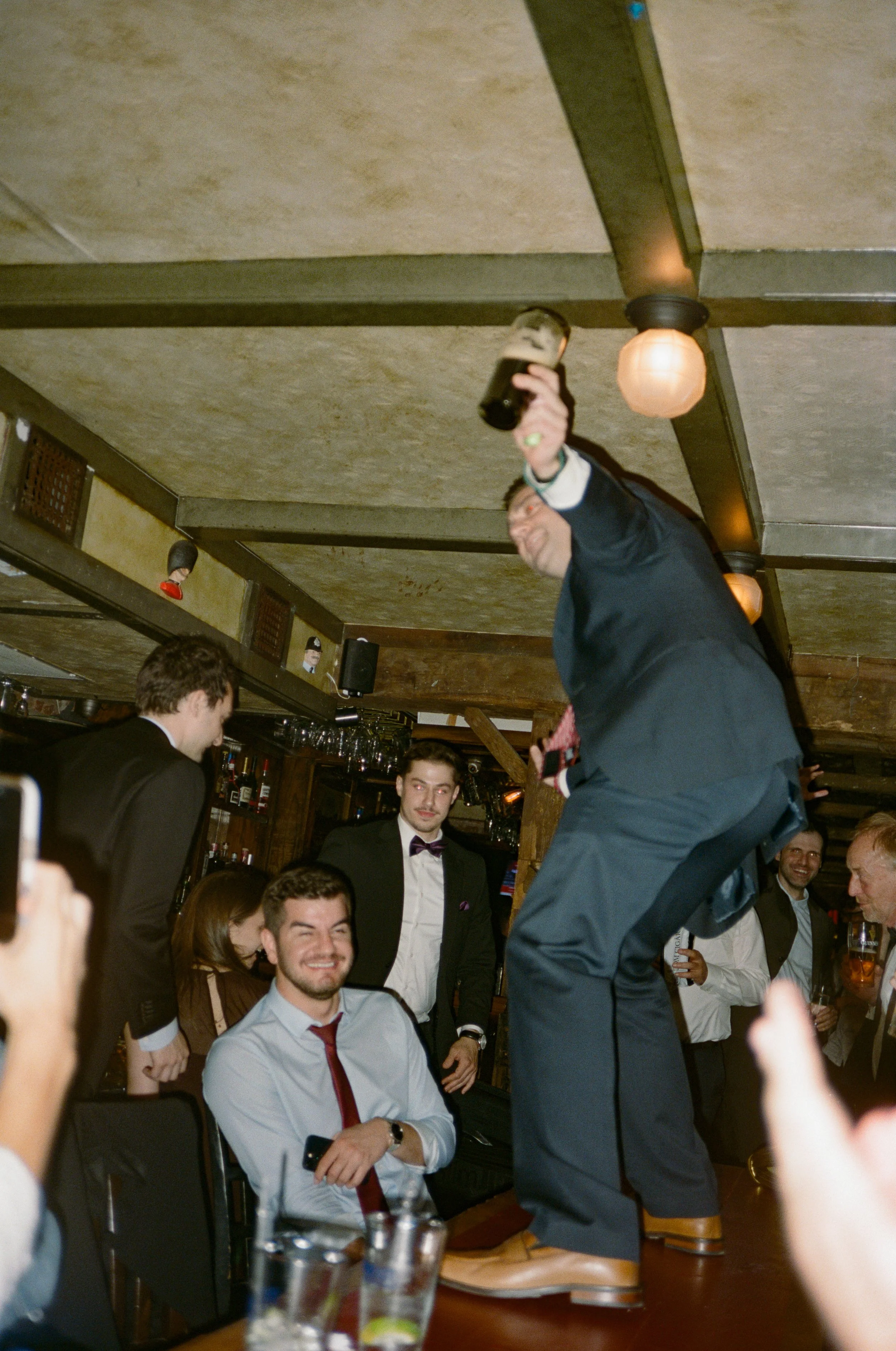 A man in a suit and bow tie standing on a table holding a drink, while other people around him cheer and smile in a lively bar scene.