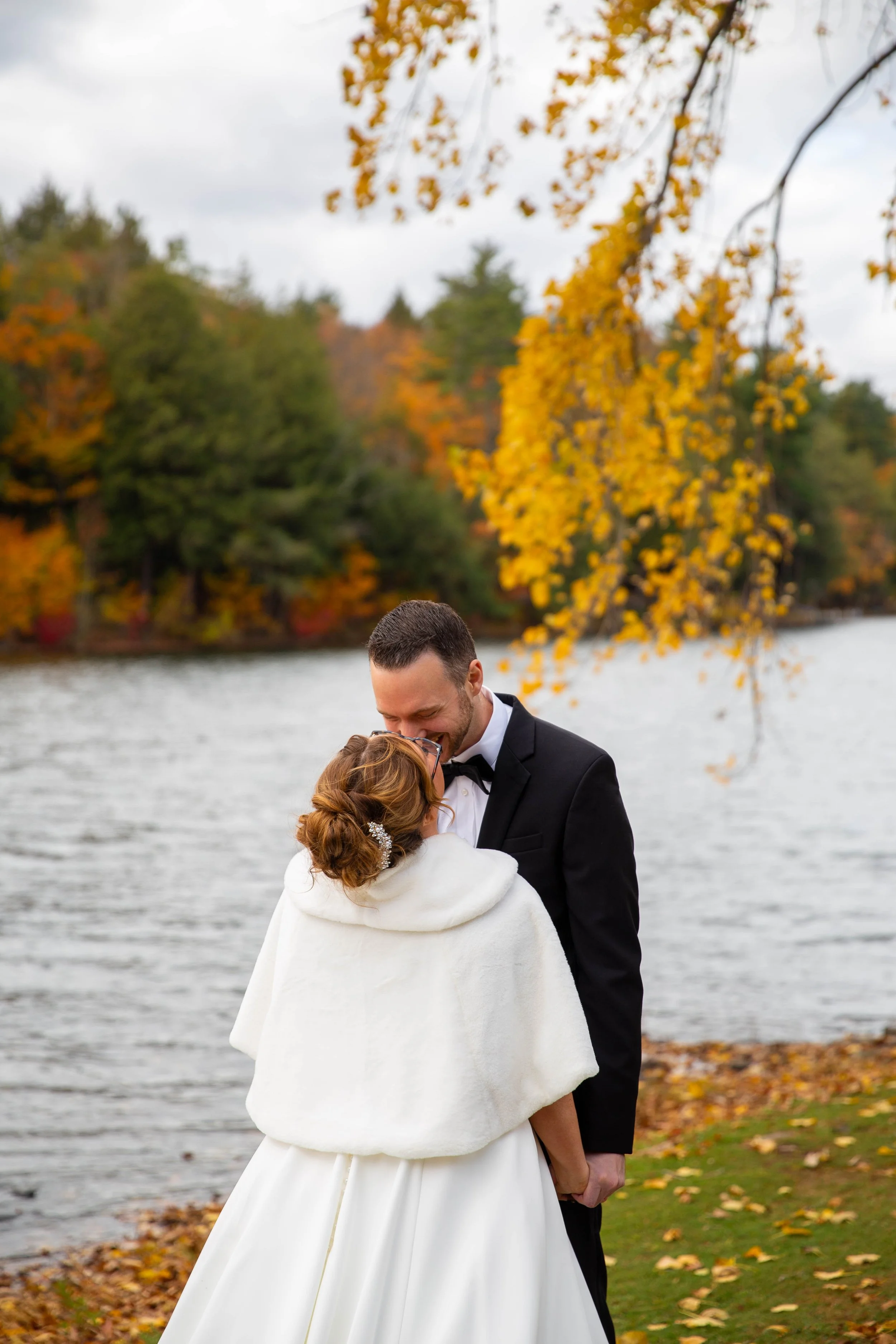 A bride and groom sharing an intimate moment outdoors near a lake during autumn, with falling leaves and colorful trees in the background.