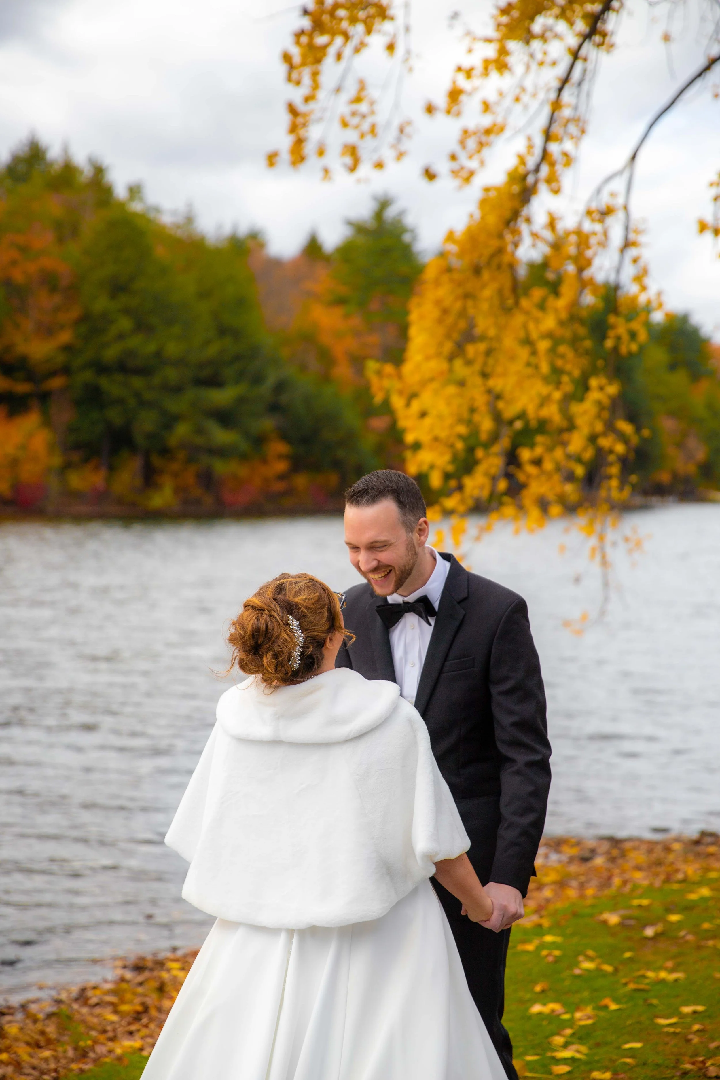 A newlywed couple holding hands and smiling at each other by a lakeside during autumn, with colorful fall foliage in the background.