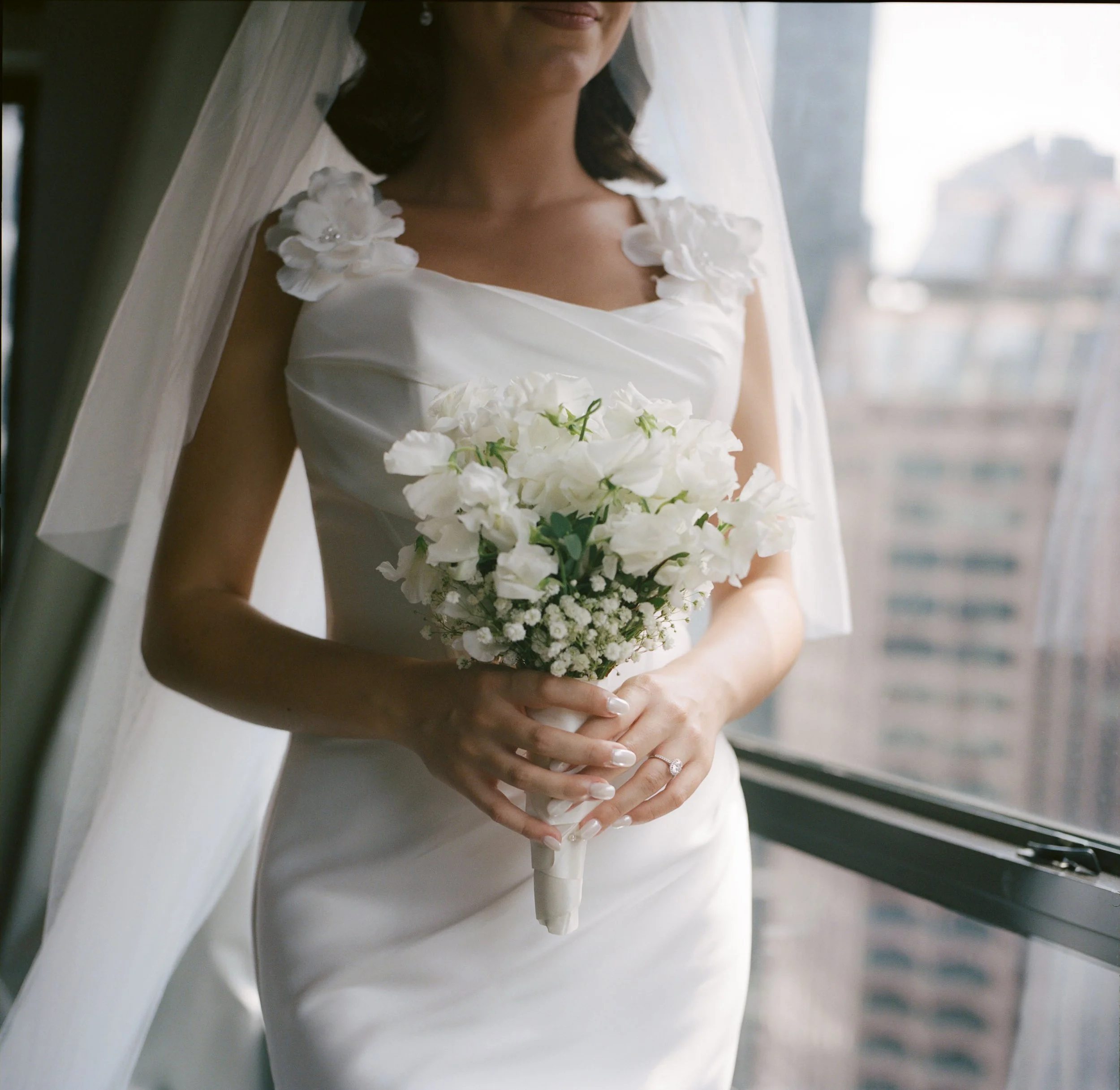 A bride holding a bouquet of white flowers, wearing a white wedding gown with floral details on the shoulders, standing indoors near a window with a city view in the background.