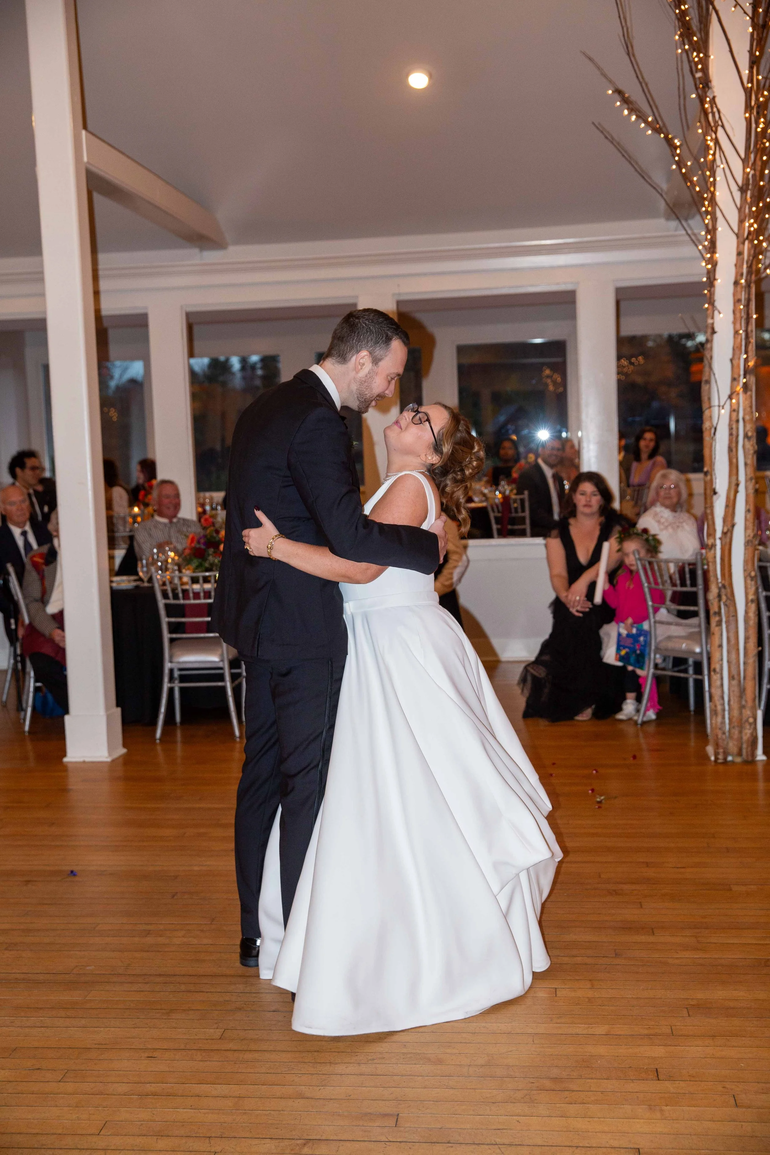 Married couple dancing at wedding reception with guests seated at tables in background and string lights decorating the venue.