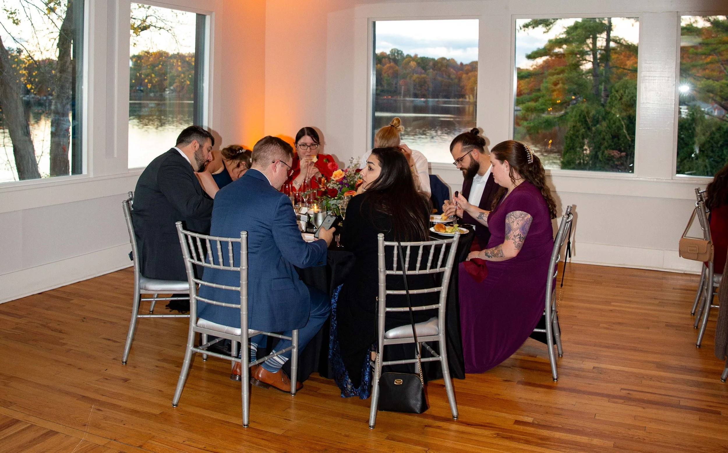 People dining at a round table with a view of a lake and trees outside the windows during evening.