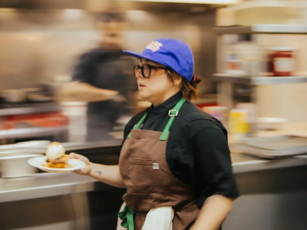 A woman in a kitchen holding a plate with a dessert topped with ice cream, wearing a blue cap, glasses, a black shirt, and a brown apron, with a blurred background of kitchen equipment.
