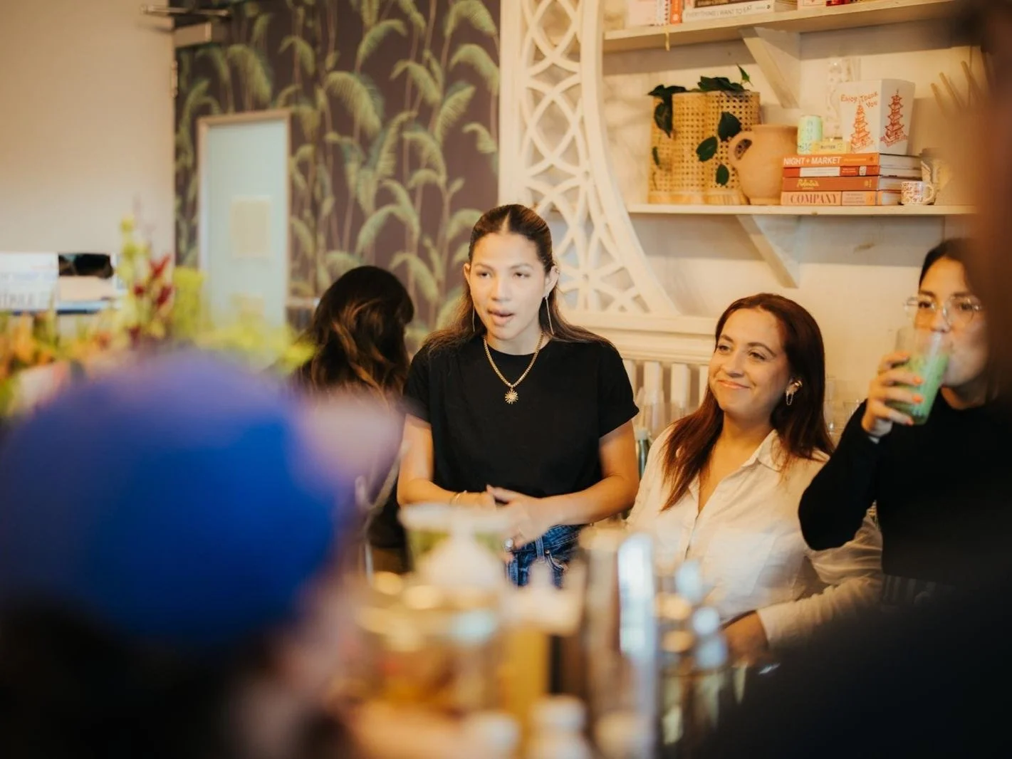 A group of women sitting at a table in a cozy café, having a conversation and enjoying drinks, with decorative shelves and plants in the background.