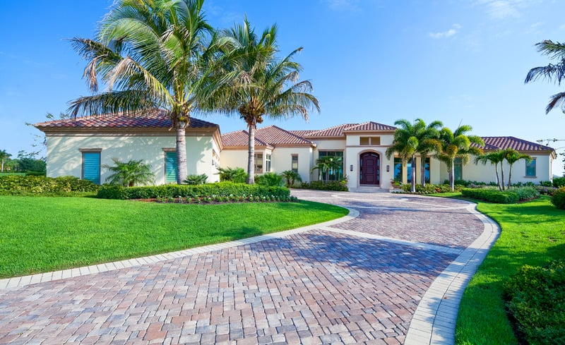 Luxury mansion with red-tiled roof, surrounded by palm trees and a green lawn, with a curved brick driveway leading to the front entrance on a sunny day.