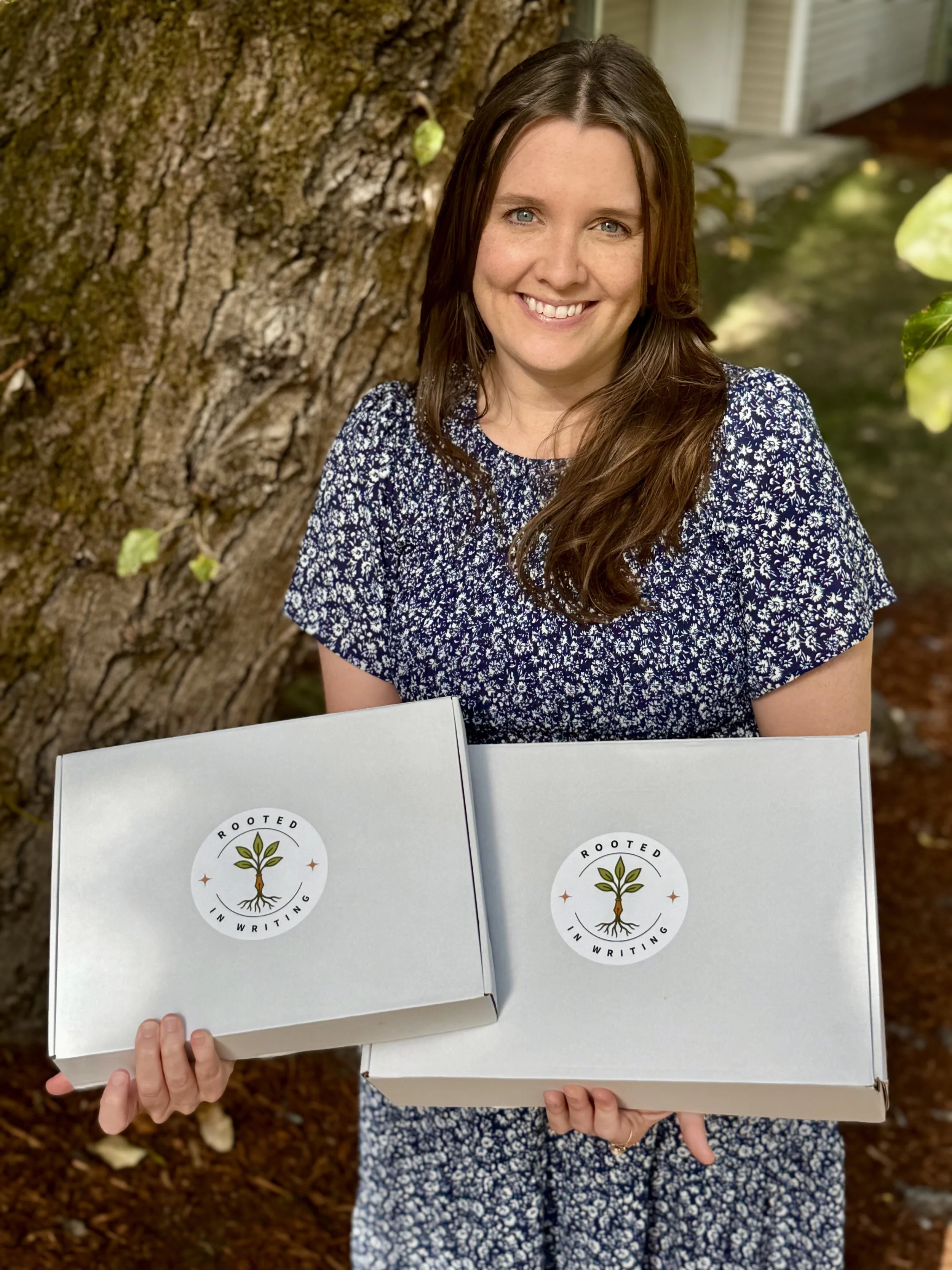 Erin Moffet, founder of Rooted in Writing, holding two subscription boxes — early literacy and fine motor skills activity kits for children ages 3–6.