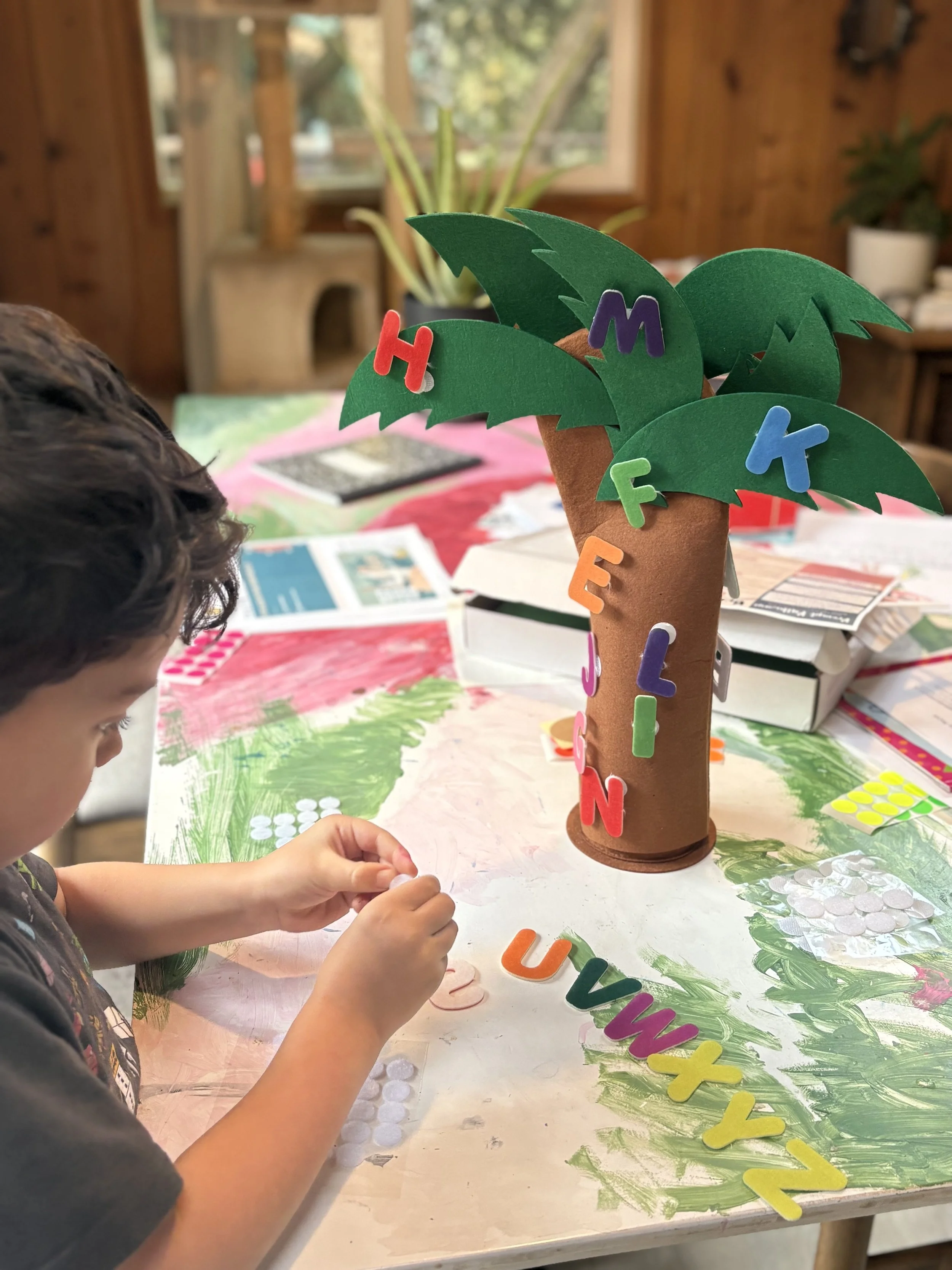 Young child assembling a colorful letter palm tree craft, a hands-on early literacy activity from the Rooted in Writing Alphabet Box for preschoolers.