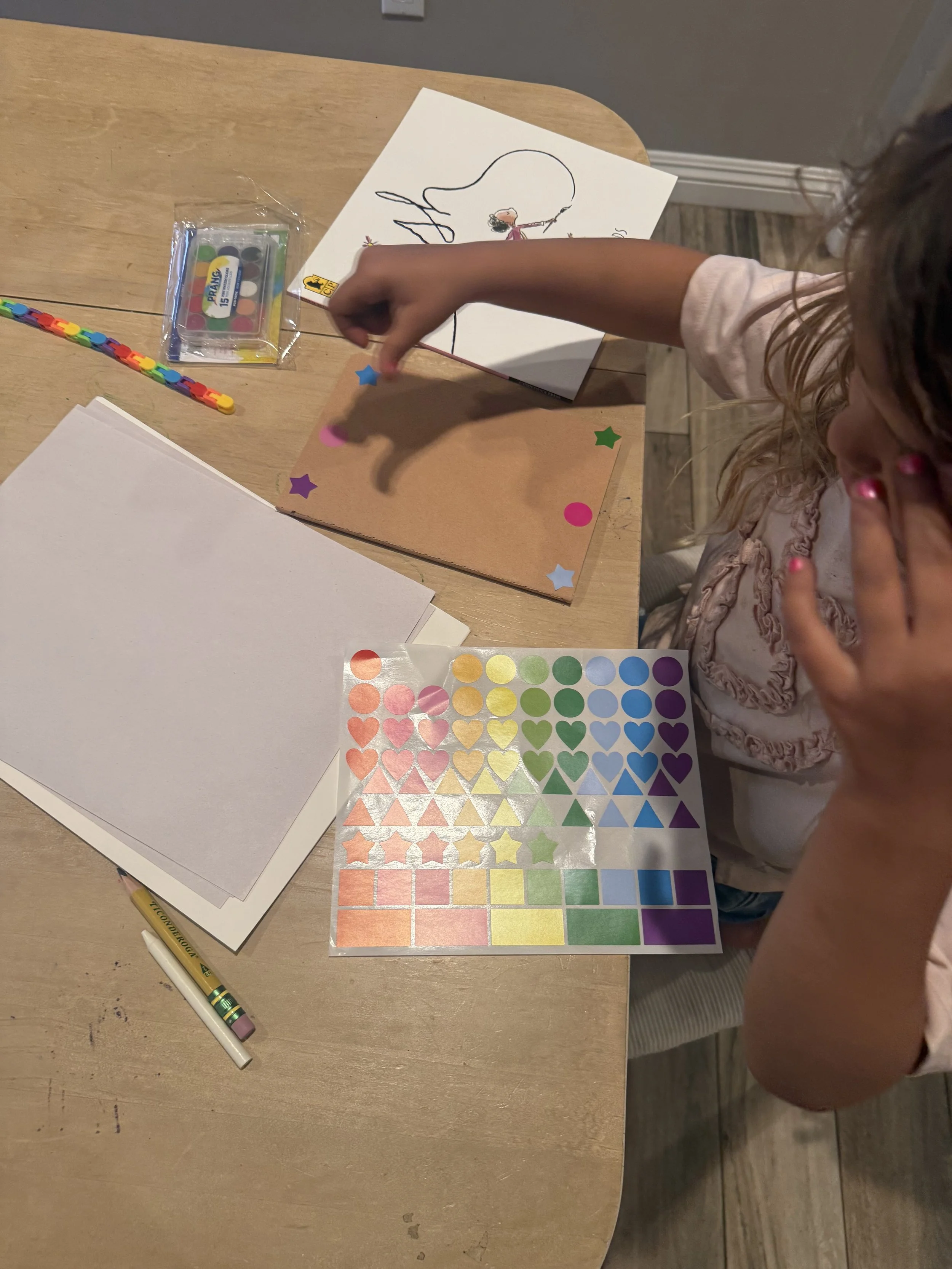 Young child placing stickers on a craft project alongside colorful shape stickers, Prang watercolors, and the book Ish — hands-on fine motor activity from a Rooted in Writing subscription box.