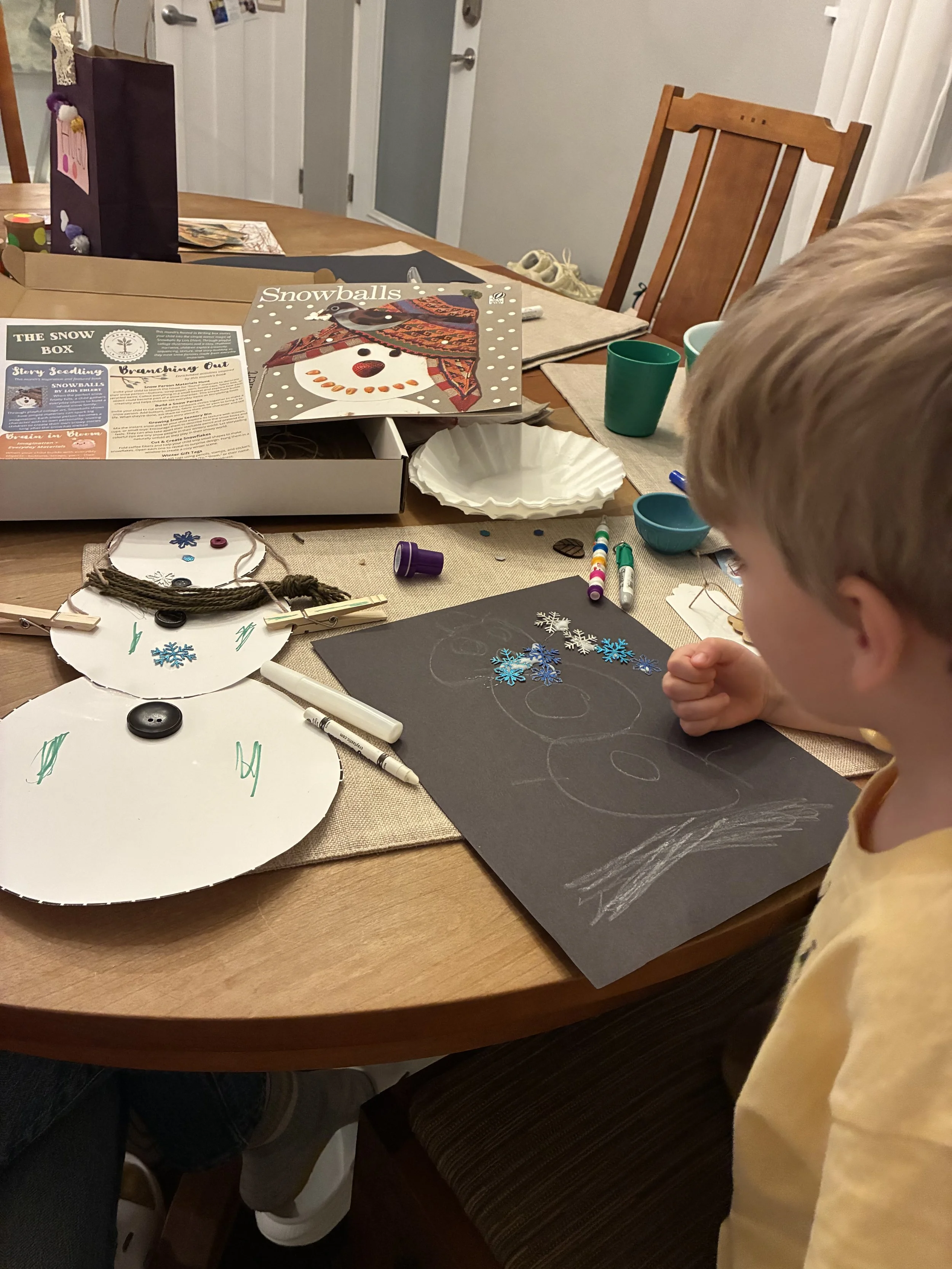 A child sitting at a table making a snowman craft with paper and decorations, with craft supplies and a snowman picture on the table.
