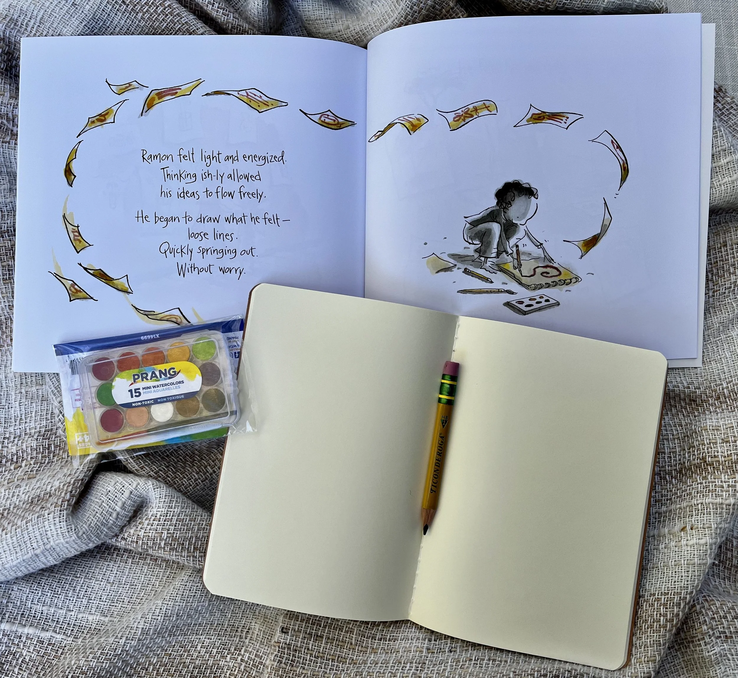 Open sketchbook with colorful illustrations of flying paper bills and a drawing of a child on the right page, surrounded by art supplies including watercolors, a crayon, and a crayon box, on a textured fabric surface.