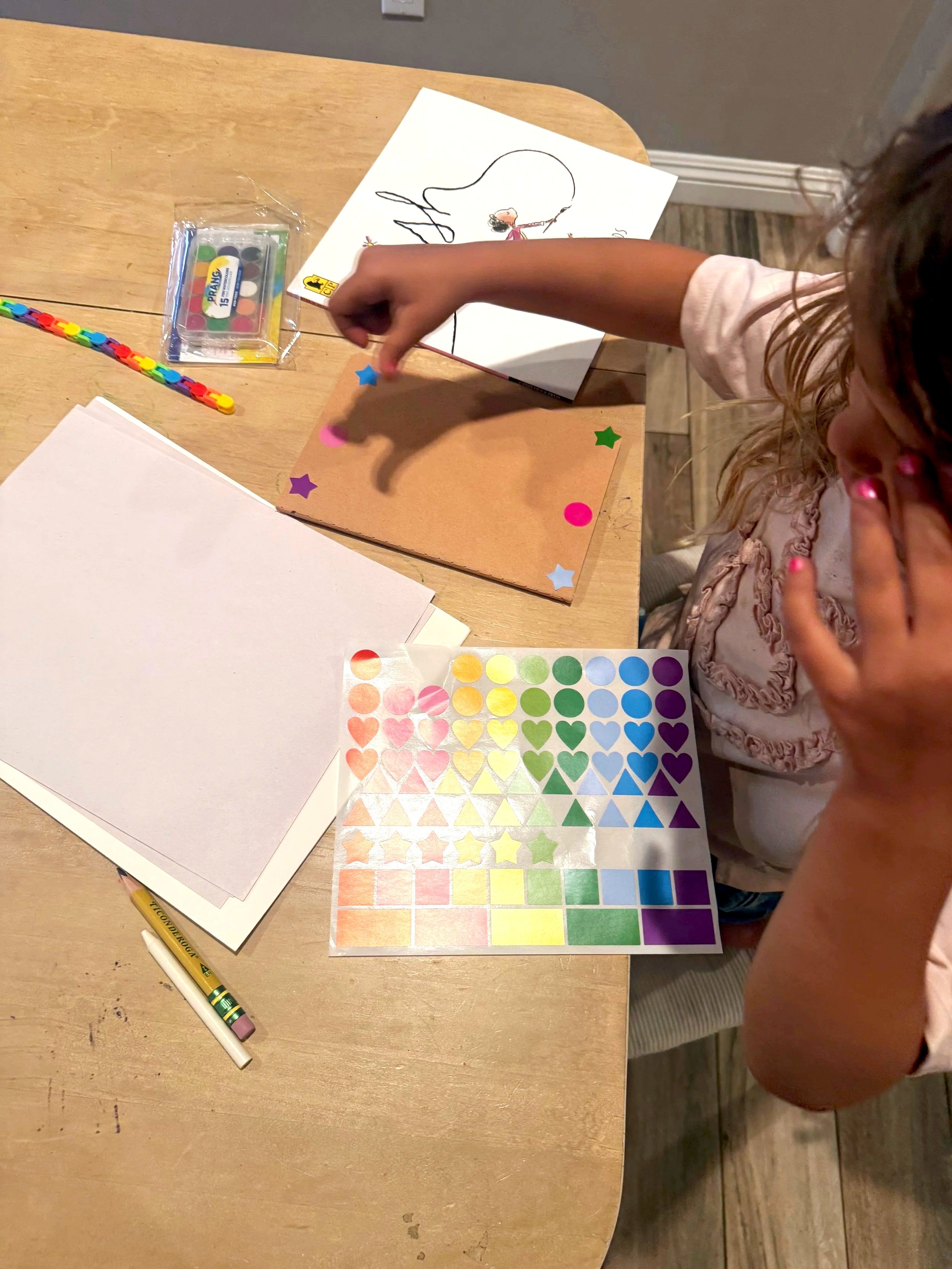 Young child placing stickers on a craft project alongside colorful shape stickers, Prang watercolors, and the book Ish — hands-on fine motor activity from a Rooted in Writing subscription box.