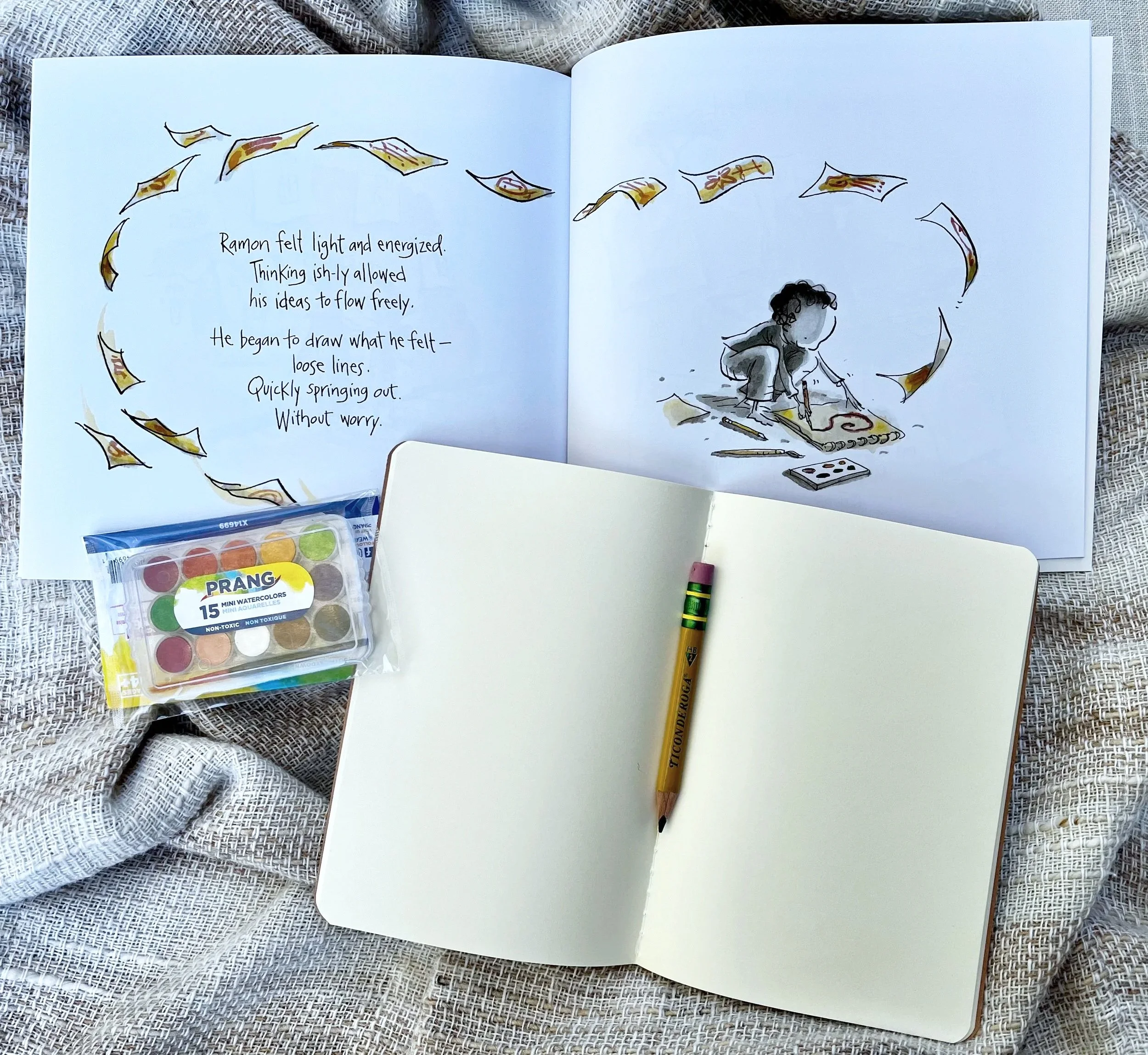 Open picture book from the Rooted in Writing subscription box beside a blank journal, Ticonderoga pencil, and Prang watercolors — hands-on early literacy materials for children ages 3–6.
