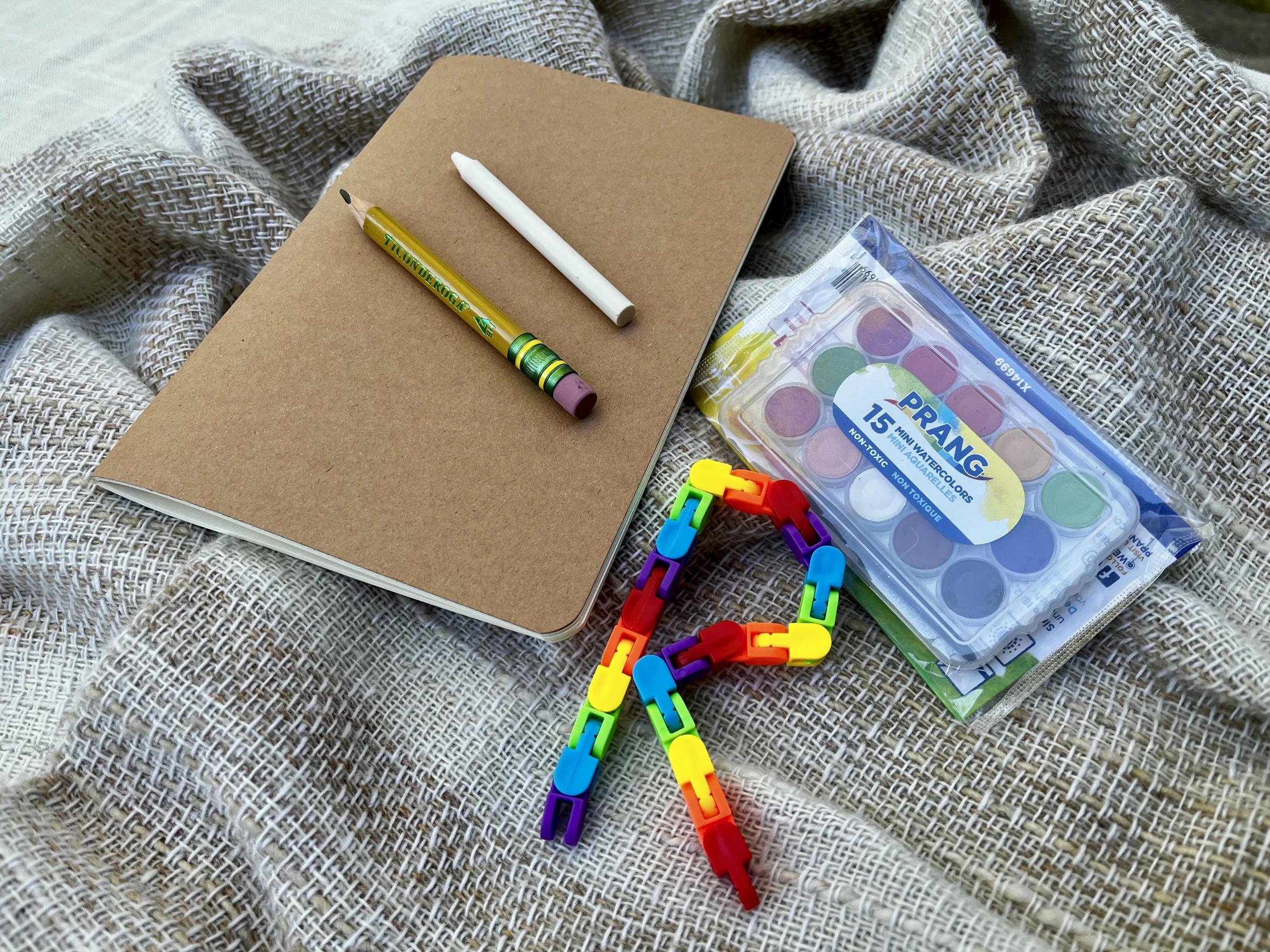 A notebook with a brown cover on a textured beige fabric, accompanied by a yellow and green marker, a white pen, watercolor paint set, colorful building blocks toy, and rainbow-colored plastic links.