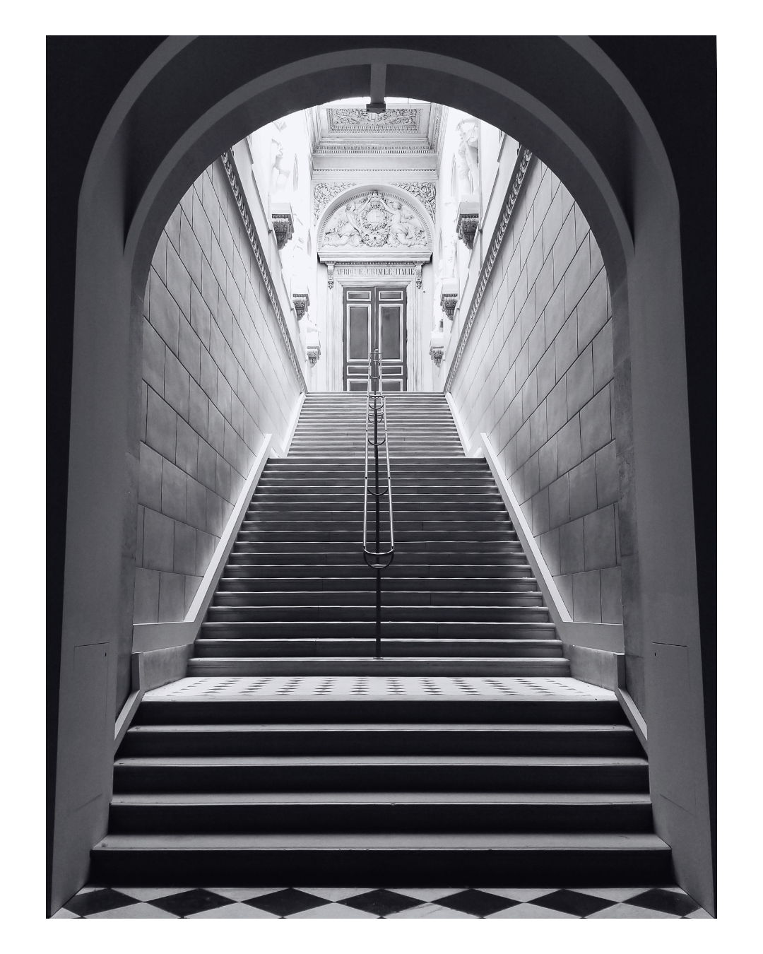 A black and white photo of a grand staircase leading to a door at the top, framed by an arched entryway with decorative walls and ceiling.