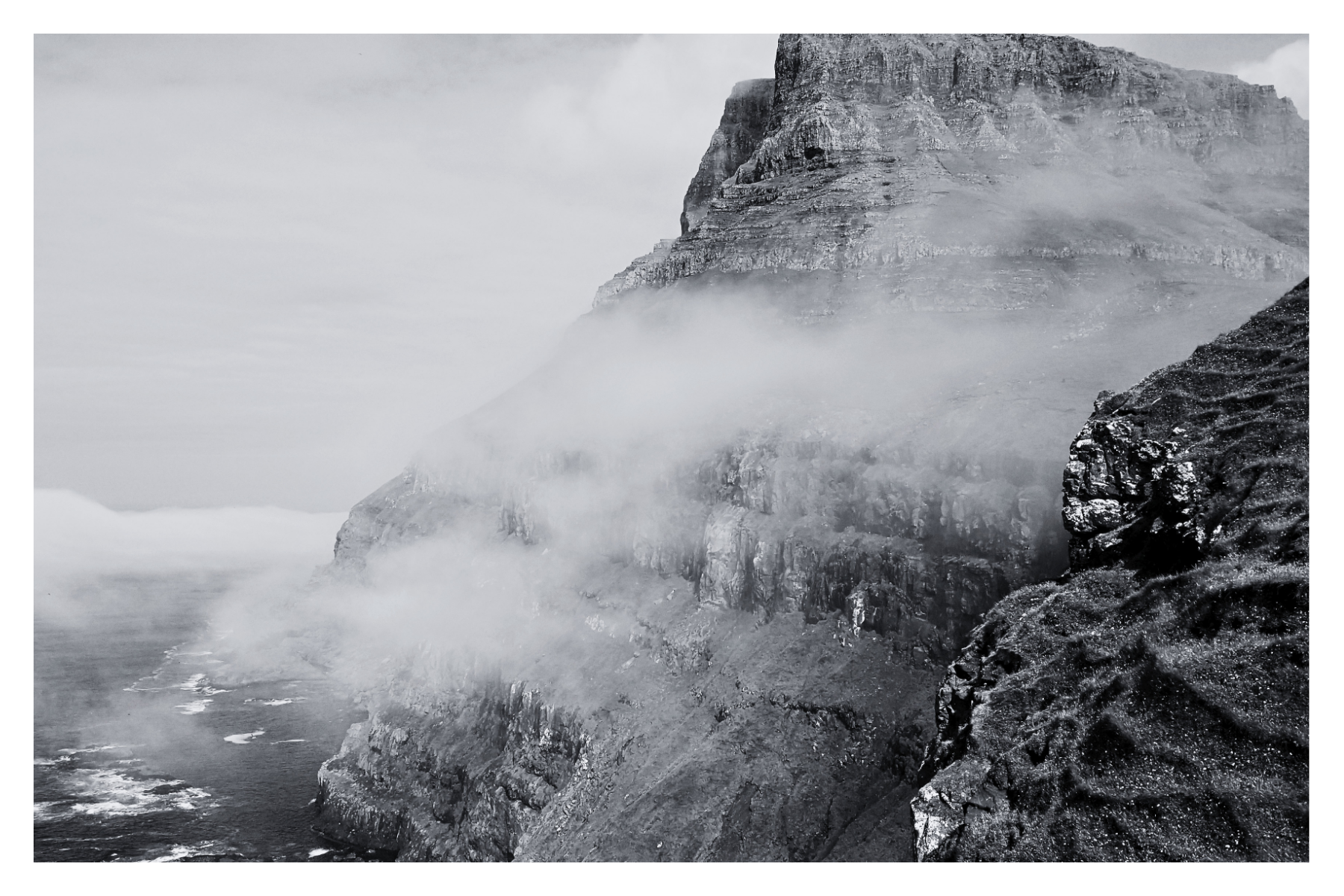 A black and white photograph of a rugged coastal cliff with waves crashing against it and mist rising from the sea.