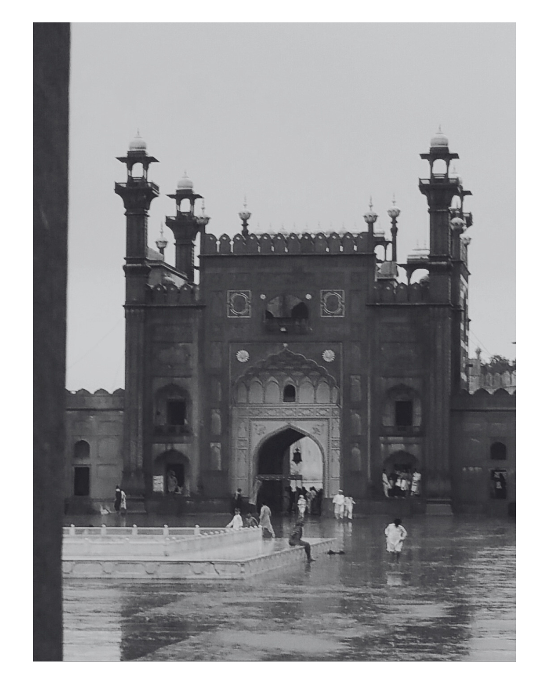 Historic mosque gate with towers, people walking, and water in the foreground.
