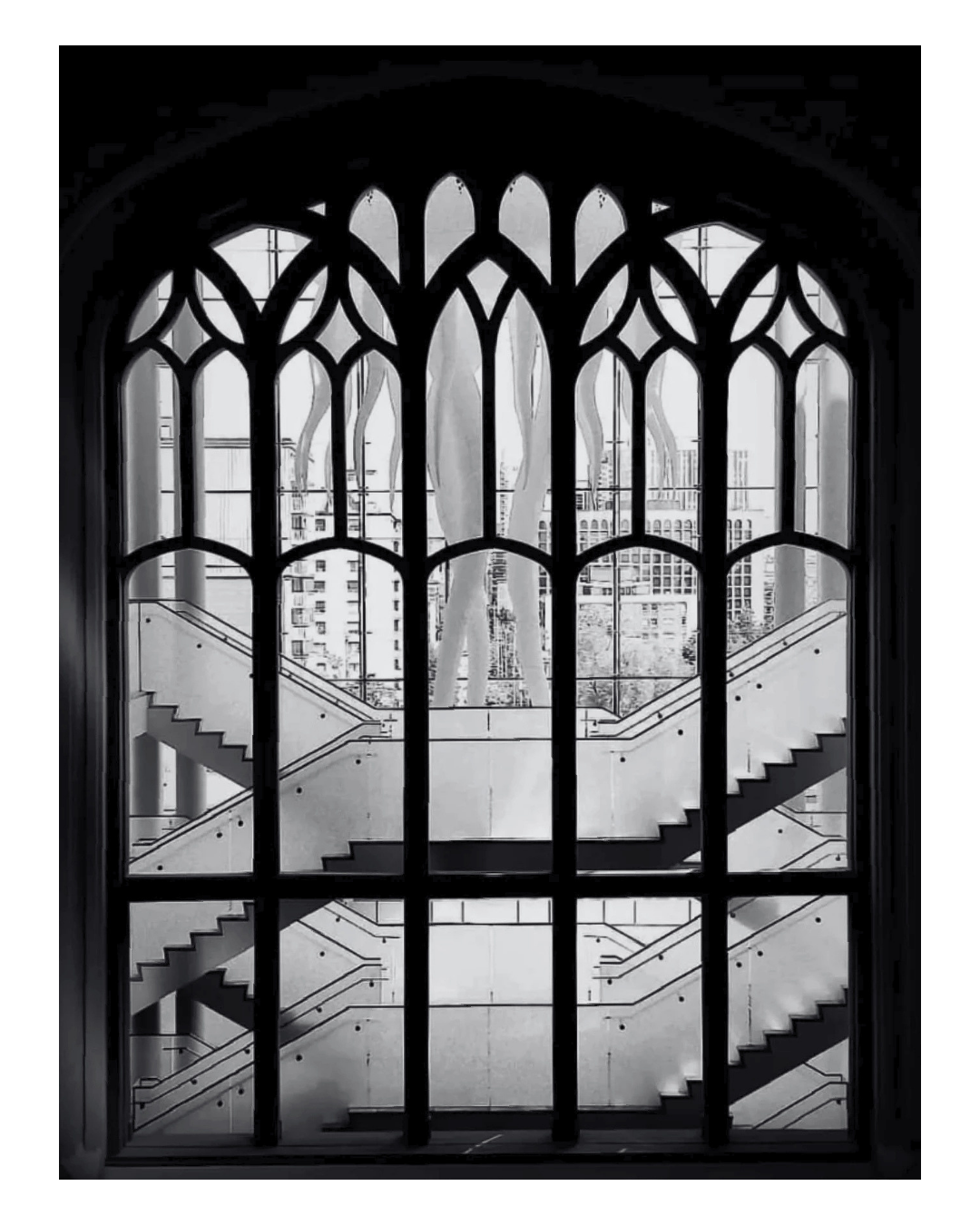 Black and white photo of an ornate arched window with stained glass design, looking out onto a modern interior space with a staircase and city buildings outside.