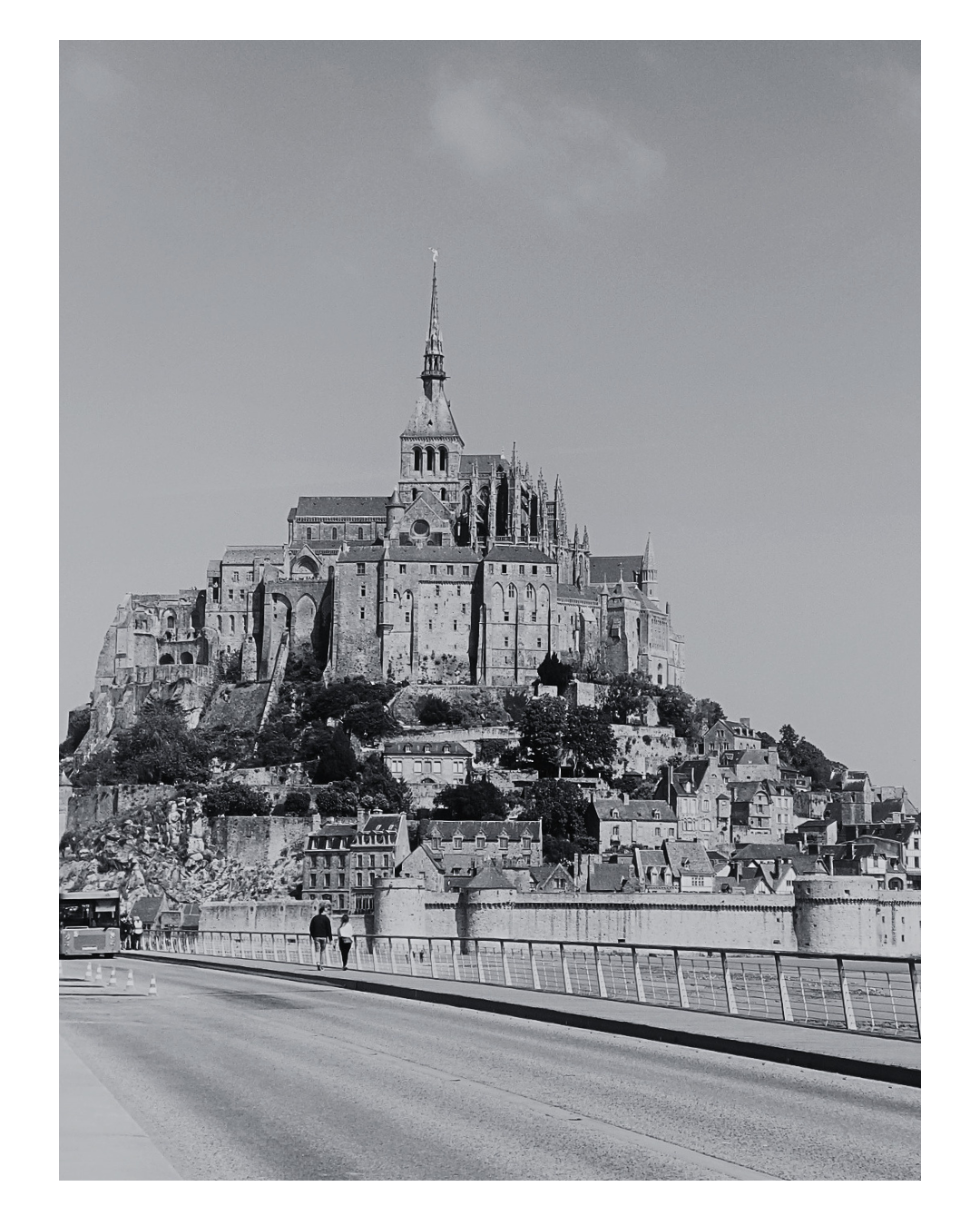 A black and white photo of Mont Saint-Michel, a historic island commune in France with a medieval abbey atop a rocky hill, surrounded by buildings and a road in the foreground.