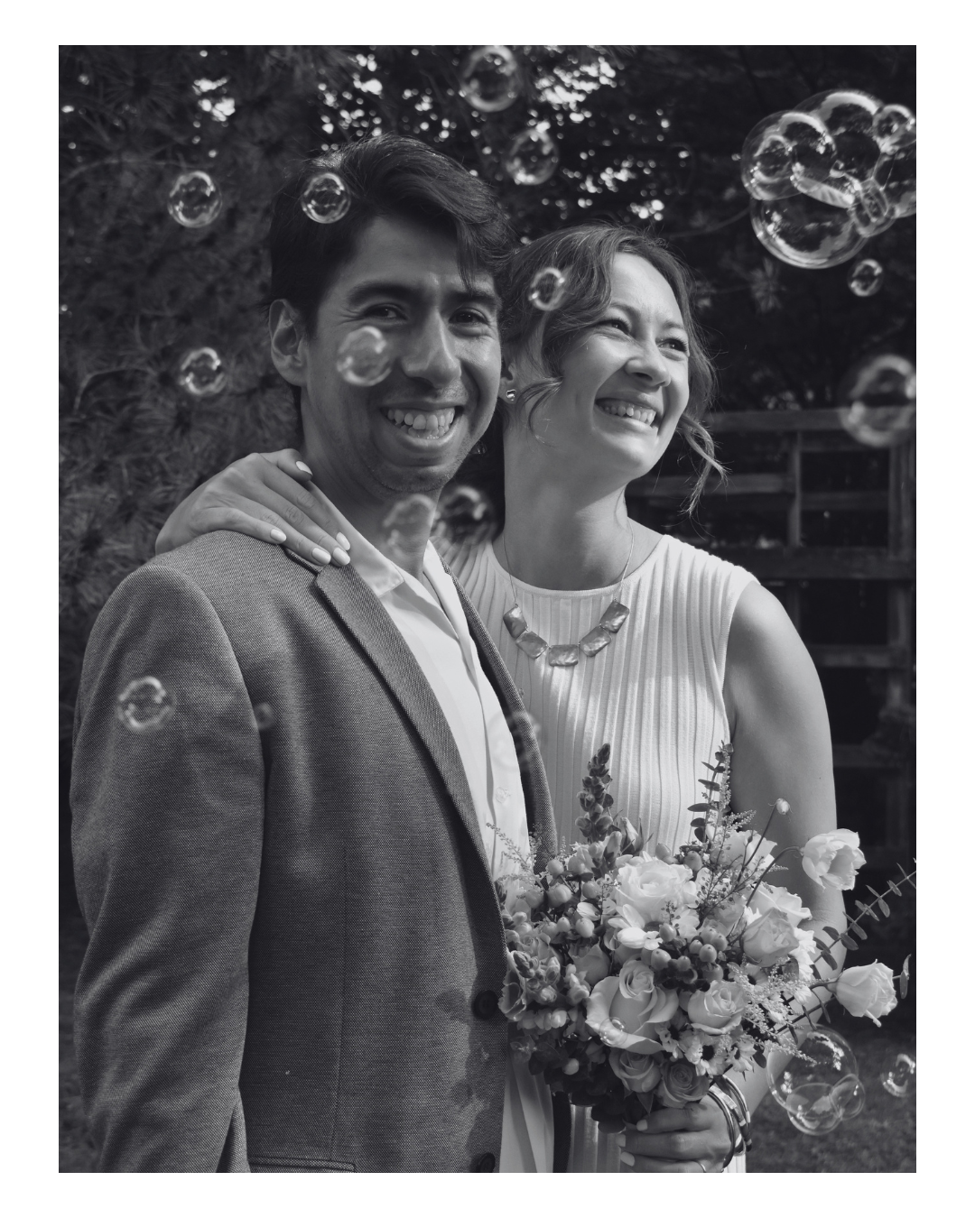 A happy couple at a wedding or celebration, with the woman holding a bouquet of flowers, surrounded by floating bubbles in an outdoor setting.