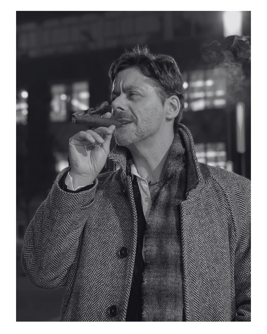 A man smoking a cigar in a black and white photo, wearing a herringbone coat and scarf, outdoors in an urban setting.