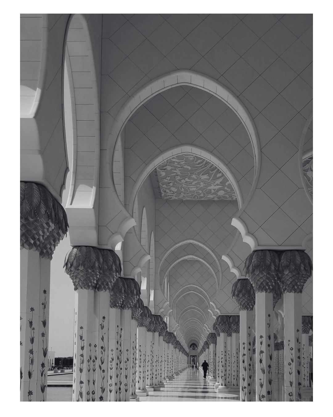 A long, decorated corridor with ornate arches and columns featuring floral patterns, leading to a person walking towards the end.