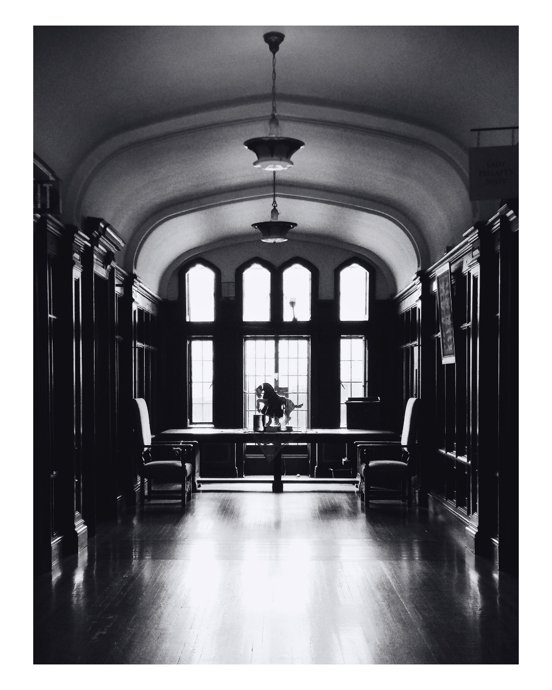 Interior of a historic wooden library or study room with chairs, large windows, and a ballerina figurine on the window sill, in black and white.